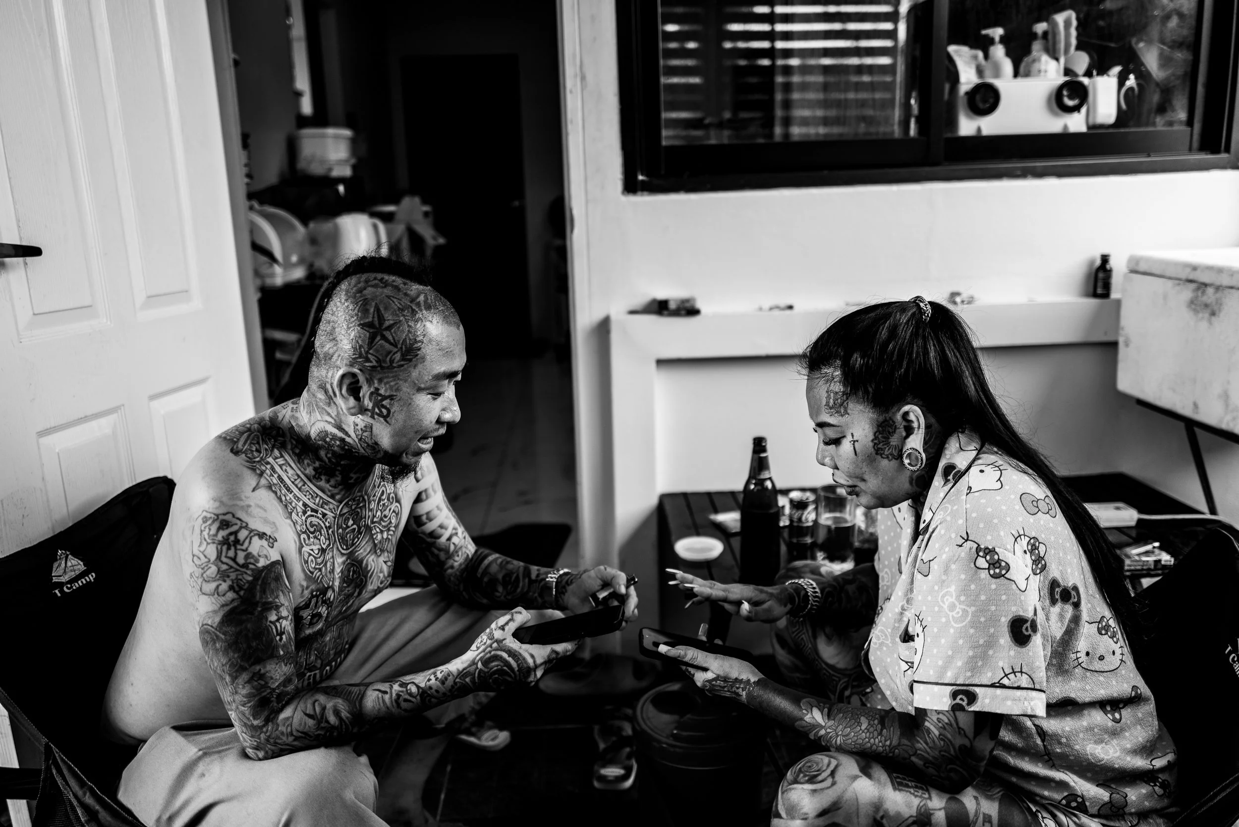 Two tattooed individuals sitting at a table, looking at their phones and smiling, in a casual indoor setting.