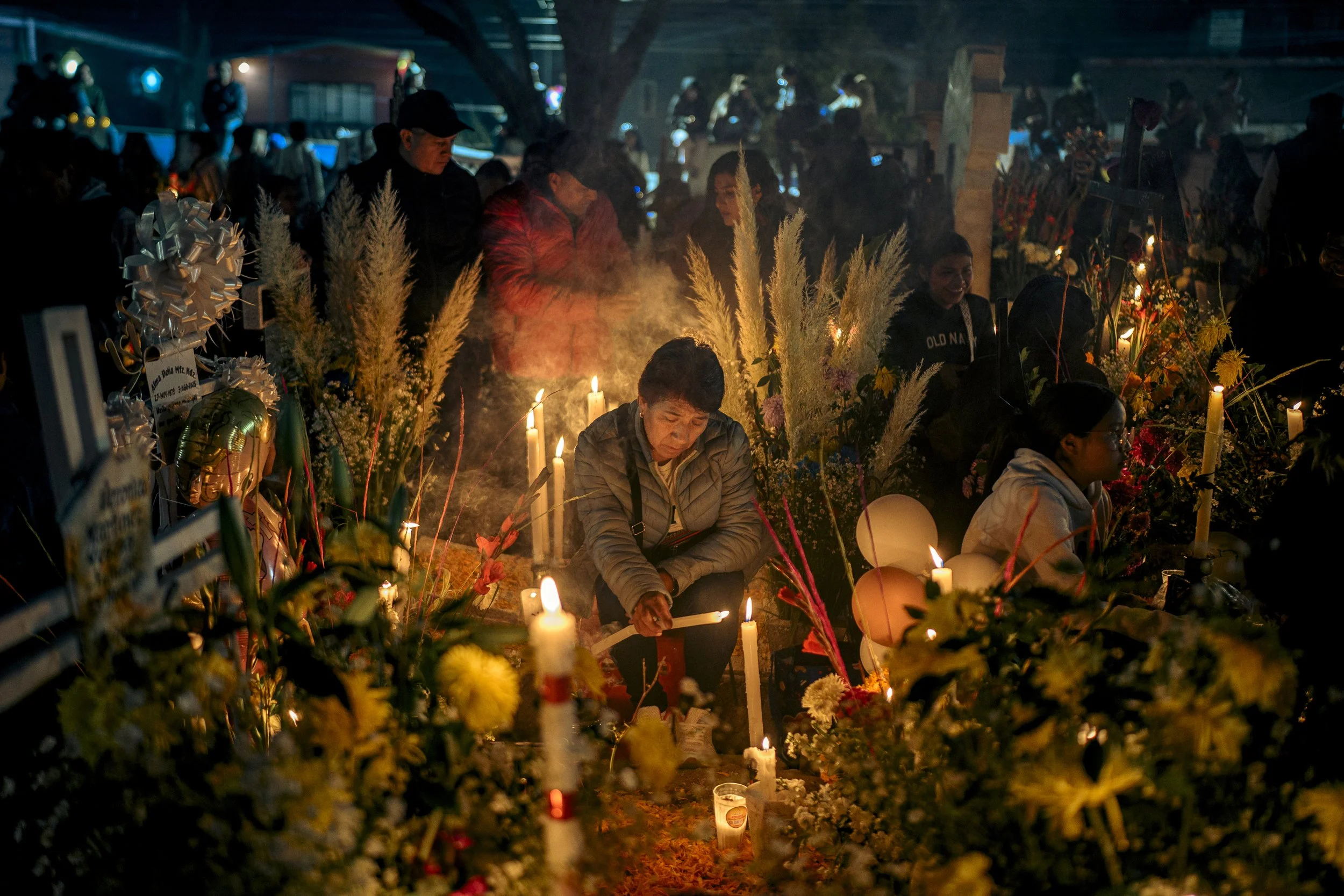 People gathered at a night memorial, lighting candles among flowers and balloons, candles emitting warm light.