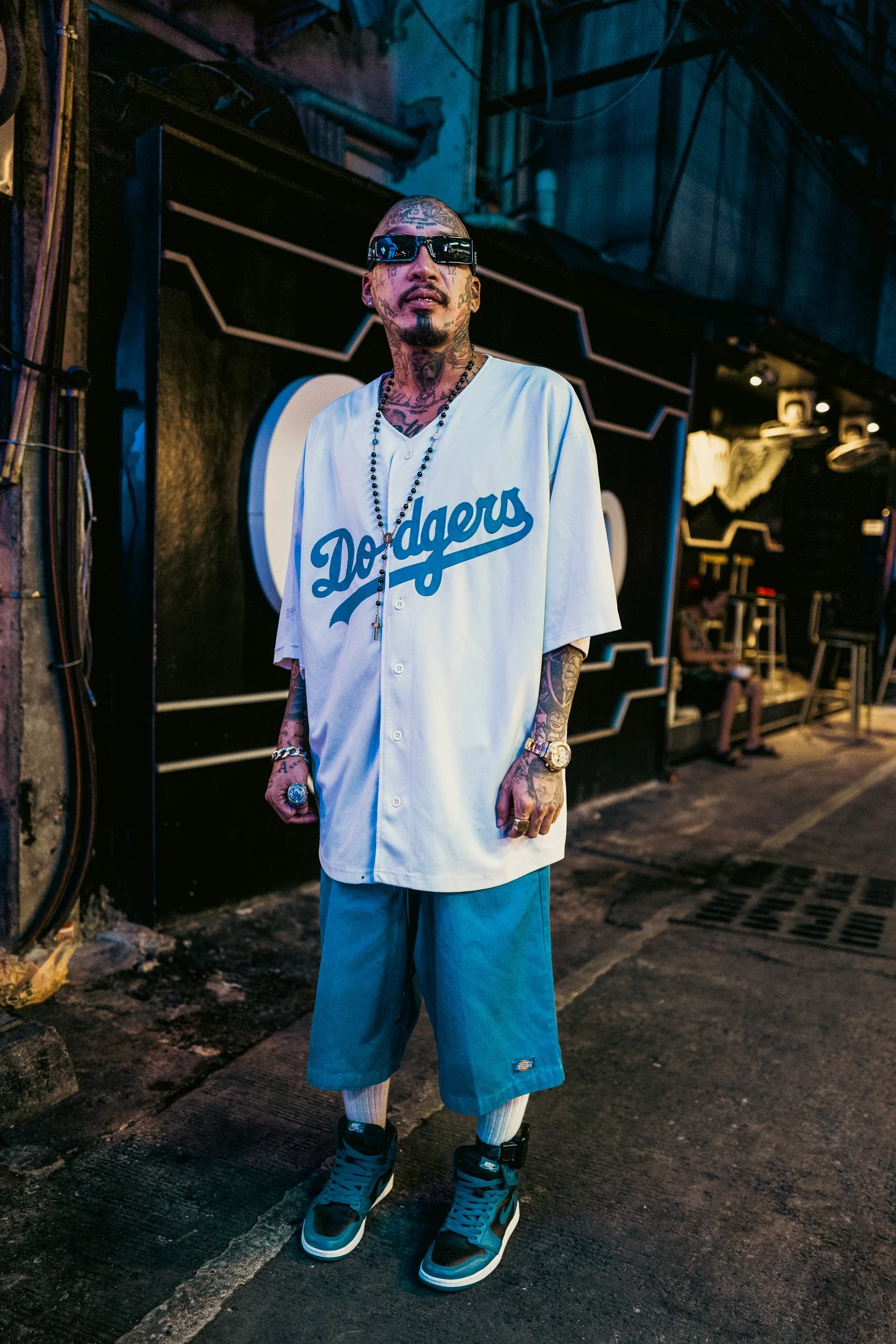 A tattooed man wearing sunglasses, a white 'Dodgers' baseball jersey, blue knee-length shorts, and black sneakers stands on a city street at night with a black and white sign behind him.