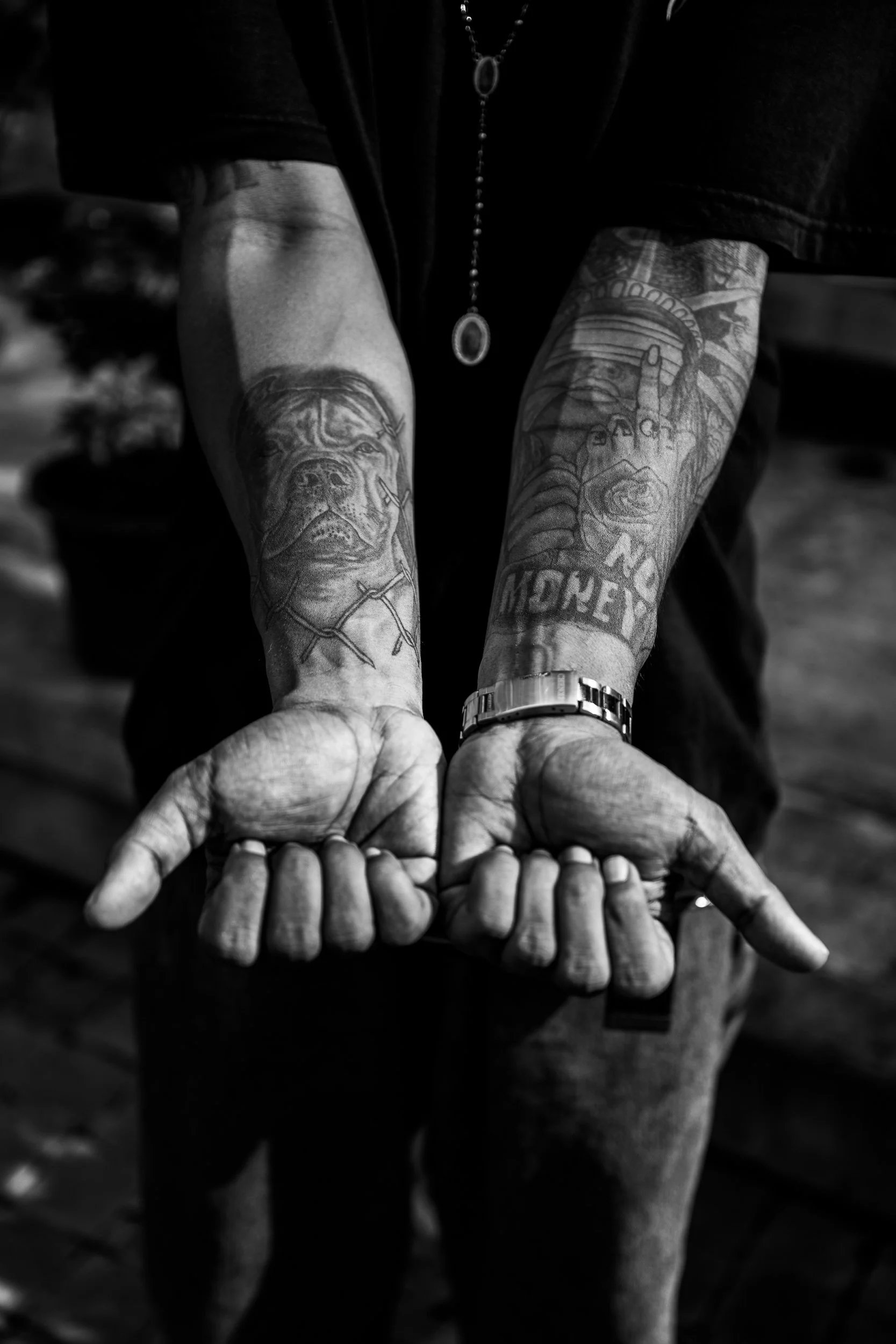 Black and white image of a person showing their forearms with tattoos, pillowing 6 fingers, wearing a watch and a necklace.
