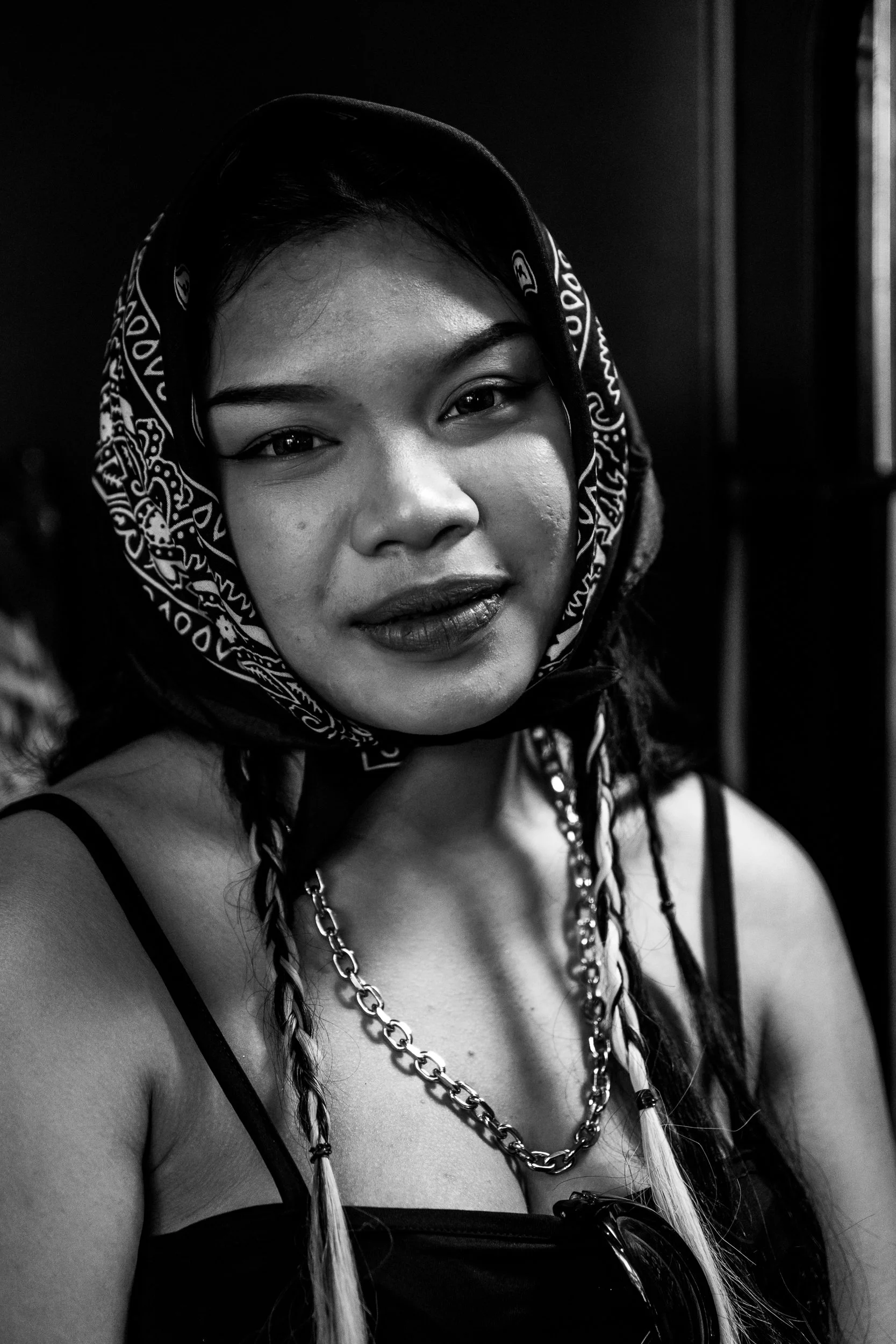 A black and white portrait of a woman with braided hair, wearing a bandana, spaghetti strap top, and layered necklaces, looking at the camera.