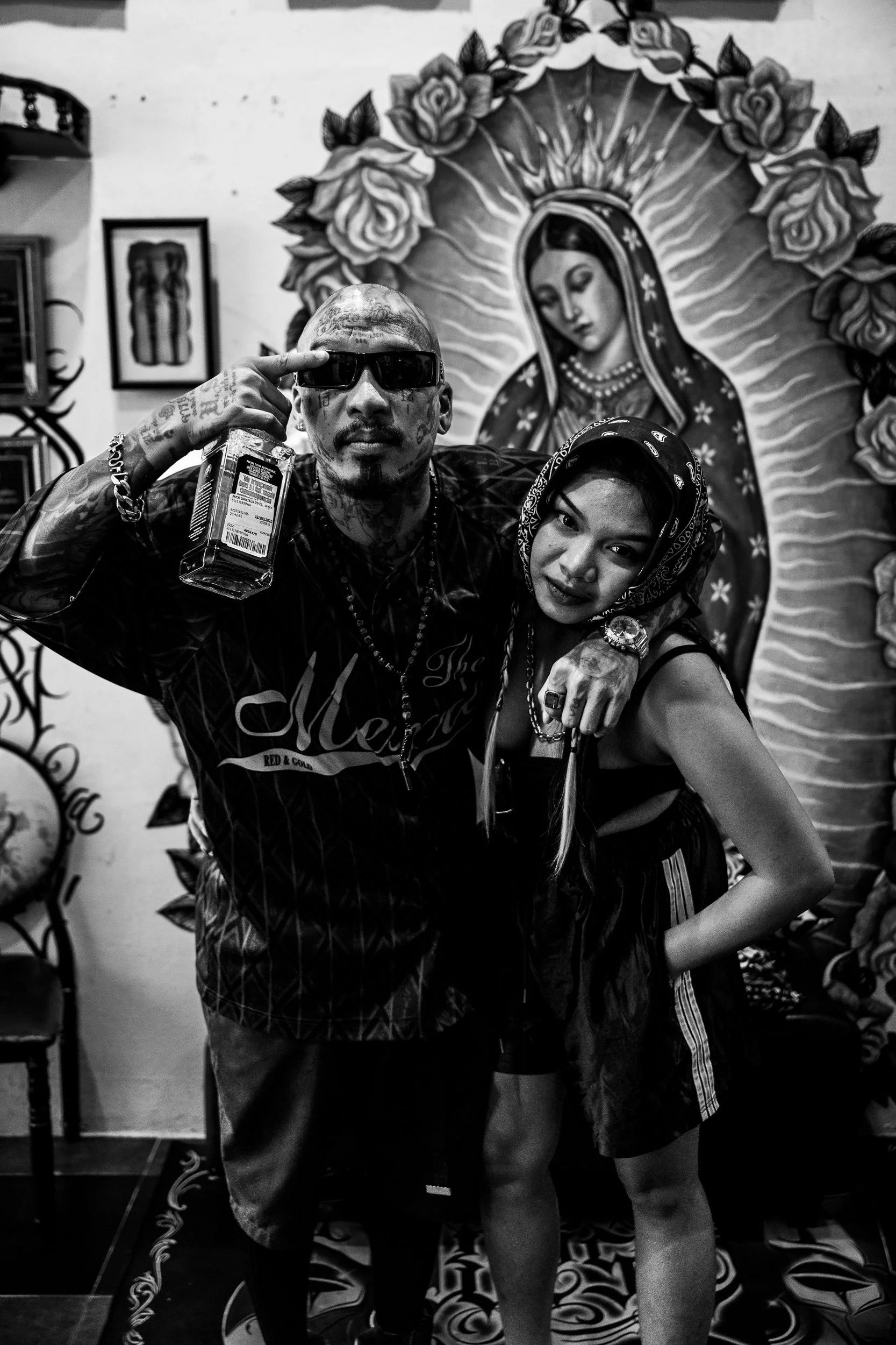 Two individuals posing in front of religious art featuring the Virgin of Guadalupe. The man on the left is wearing sunglasses, tattoos, and a patterned shirt, holding a drink, with his arm around the woman's shoulder. The woman on the right is wearin