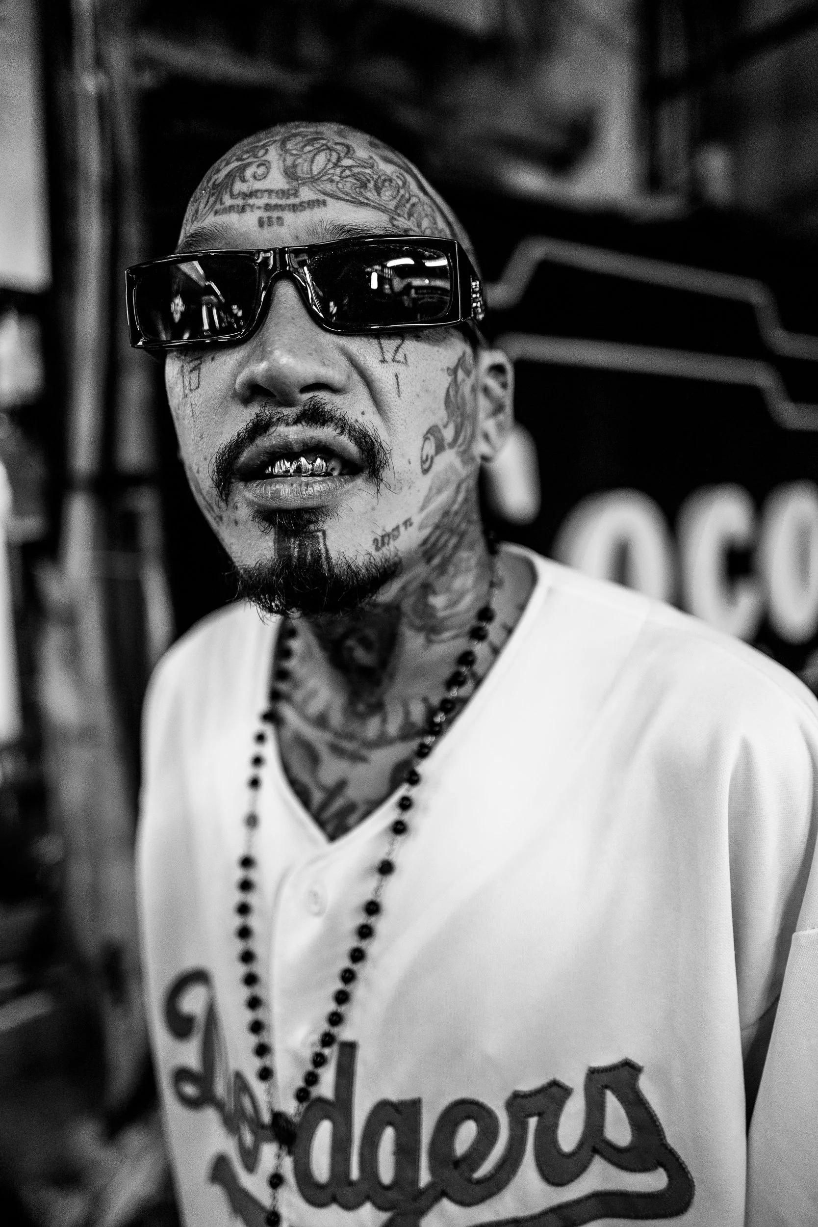 A man with tattoos covering his face and head, wearing sunglasses, gold teeth, a white shirt with text, and a beaded necklace, in a black and white close-up portrait.