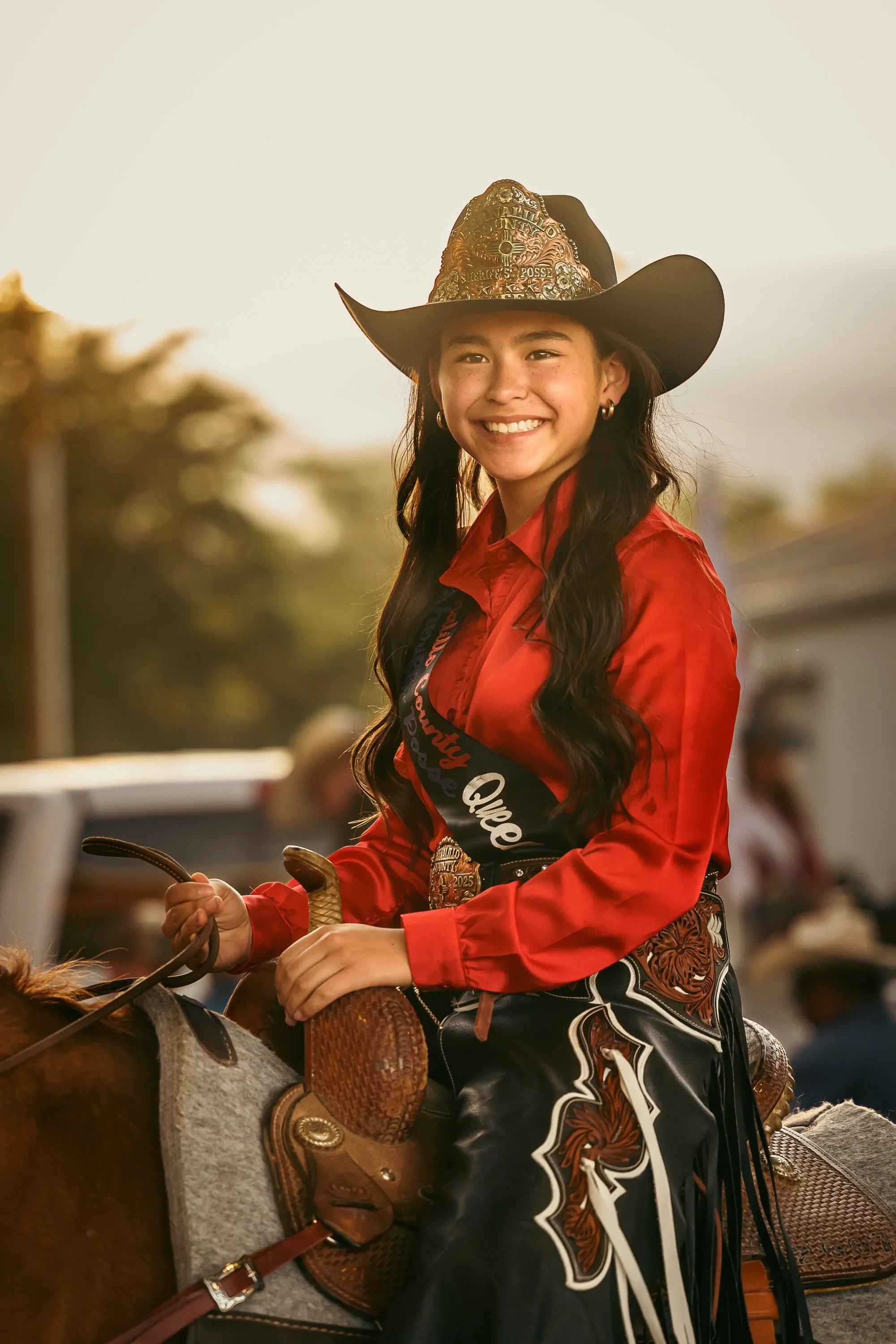 A young woman smiling and wearing a cowboy hat, red shirt, and decorative western-style pants, sitting on a horse during a rodeo event.