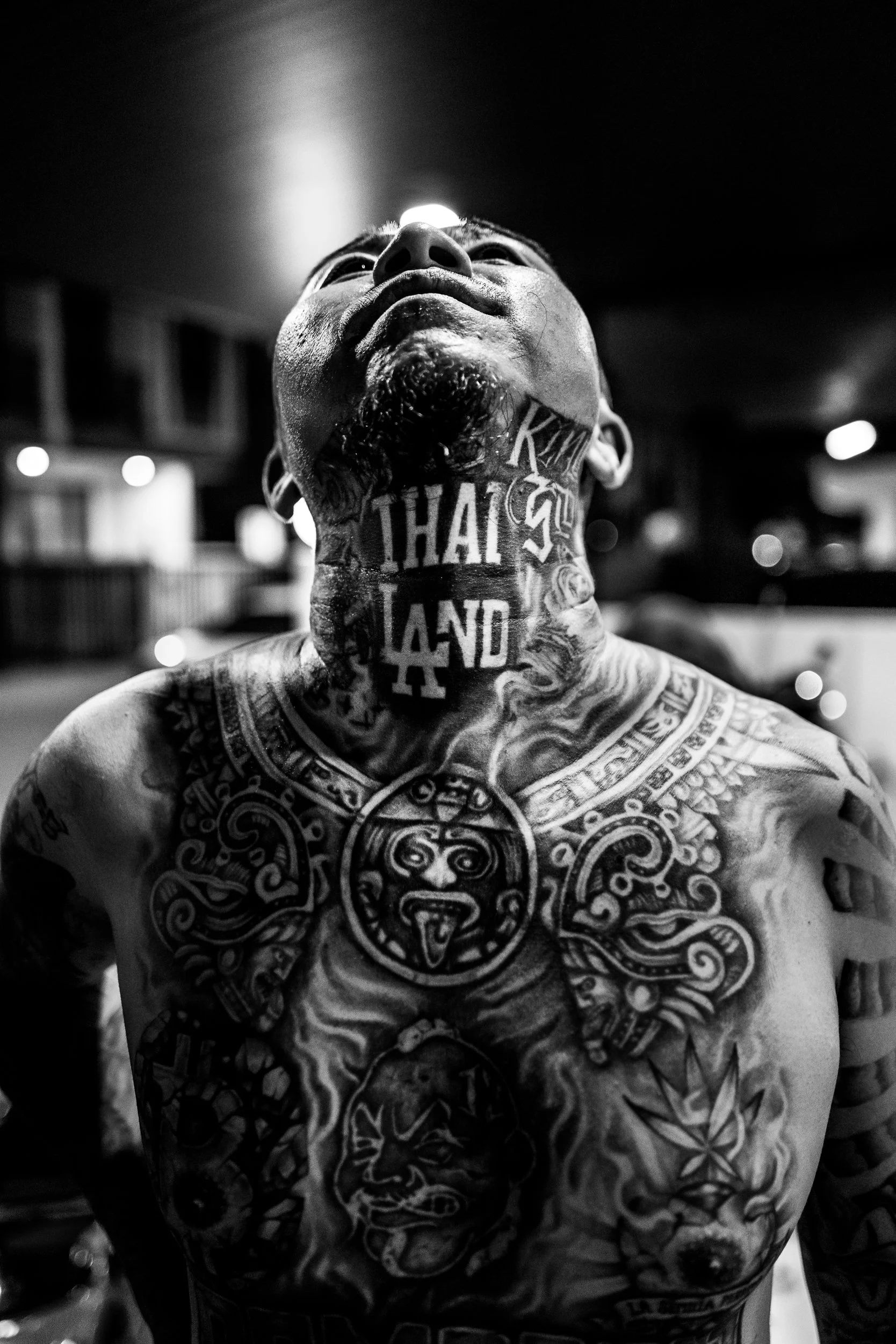 A black and white photo of a shirtless man with multiple tattoos on his chest and neck, looking upward with his head tilted back.