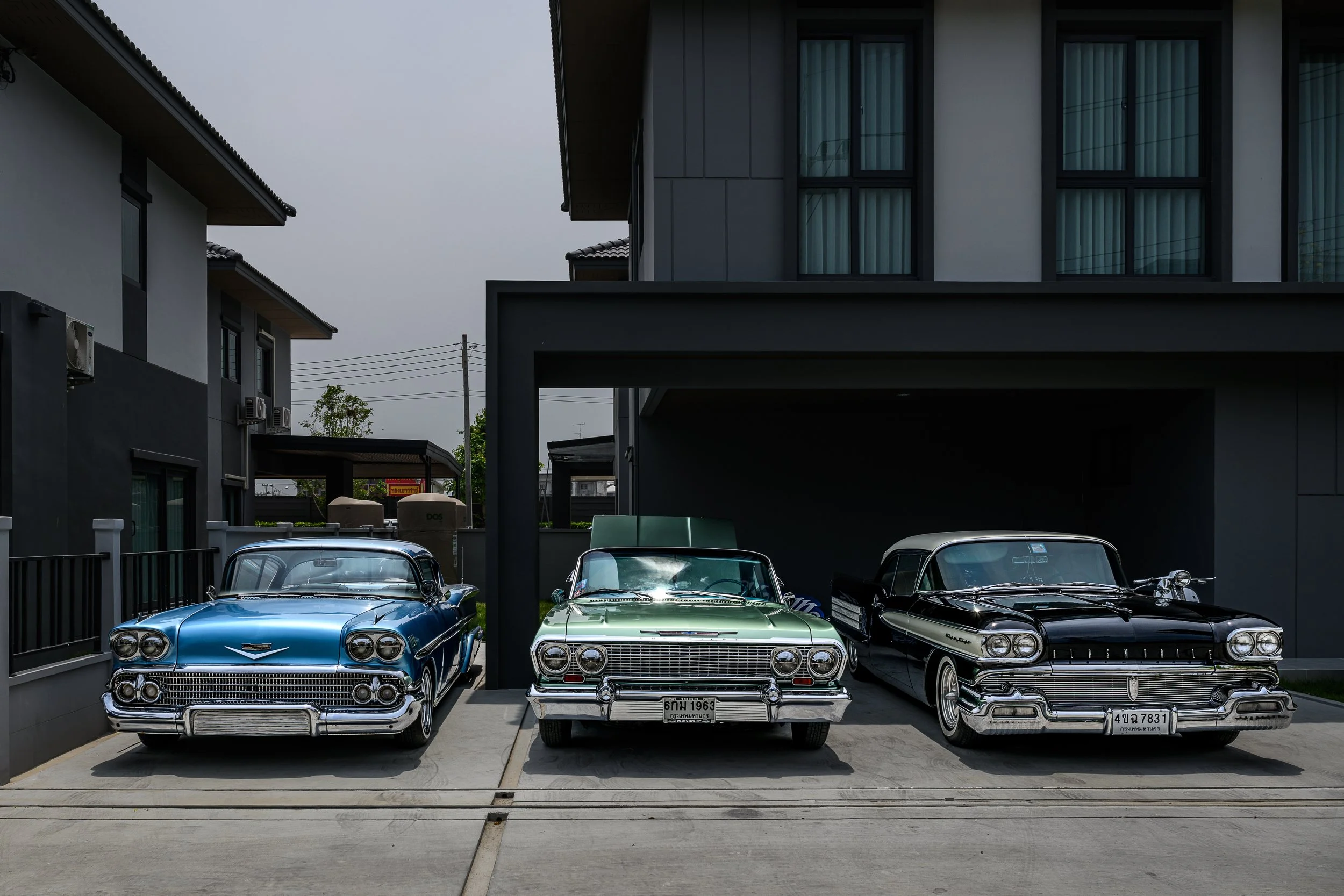 Three vintage classic cars parked in a driveway in front of a modern house, with the middle car being a green 1963 Buick.