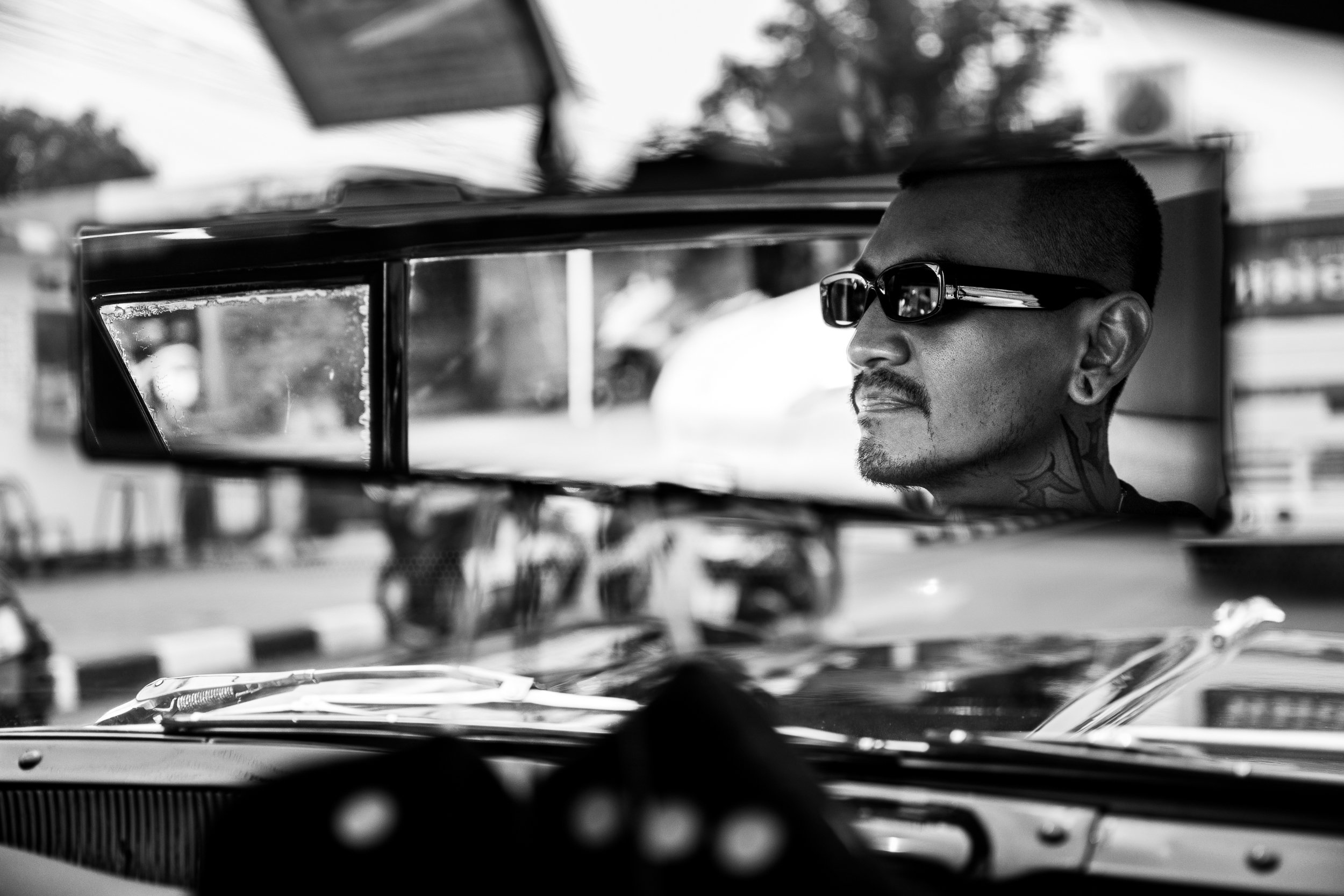 A man with short hair, wearing dark sunglasses, and sporting a tattoo on his neck, sitting in a vintage car, with the rearview mirror showing his profile.