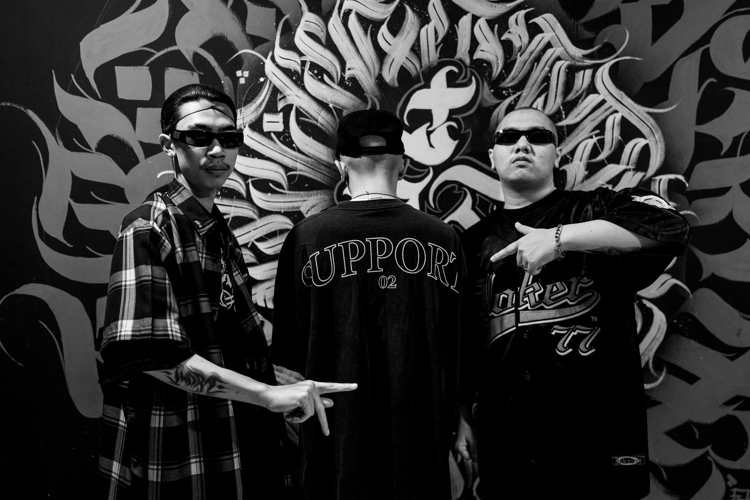 Three men wearing sunglasses and streetwear posing in front of a graffiti mural, with one pointing and another making a gesture, in a black-and-white photo.