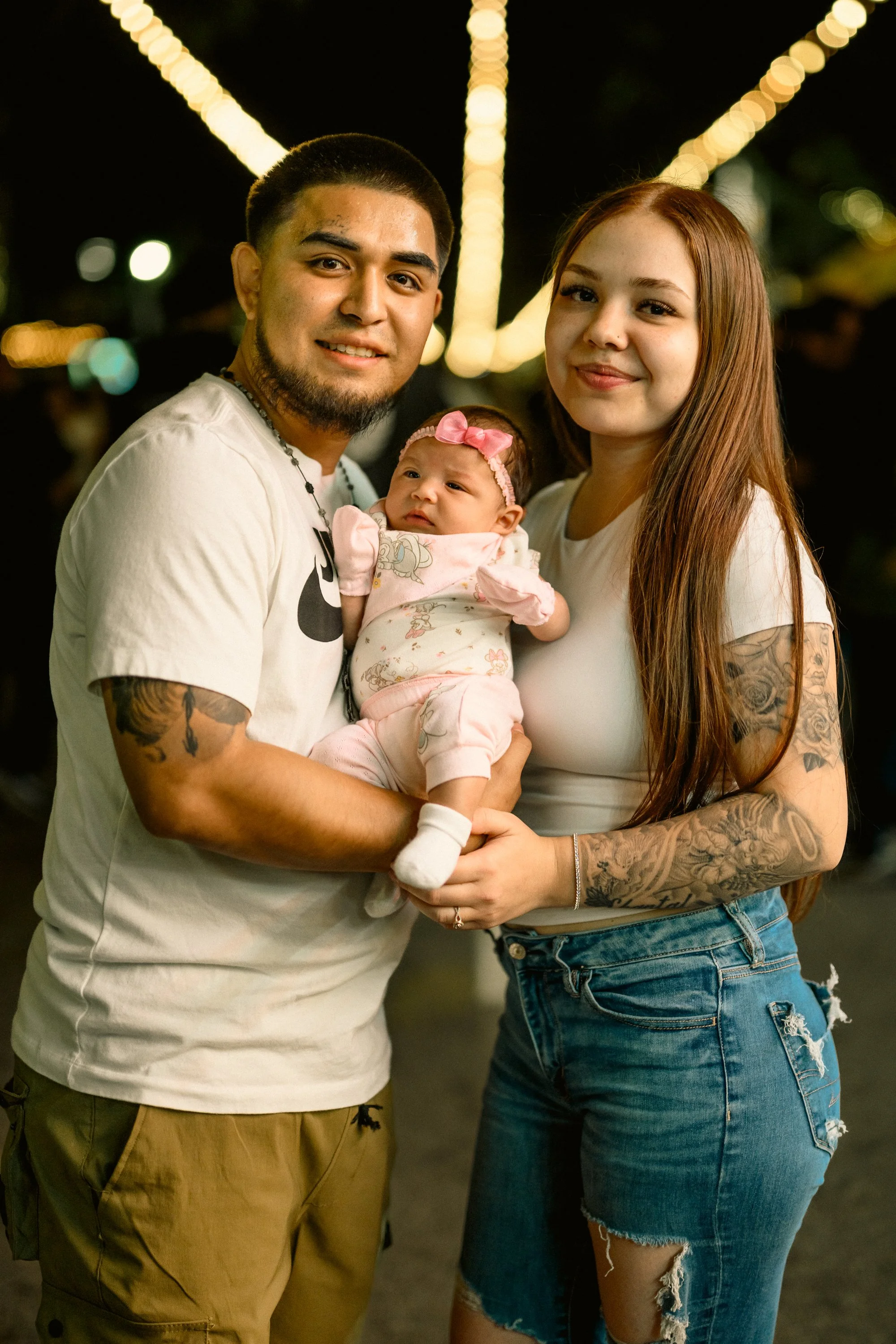 A young family posing together outdoors at night, with string lights in the background. The man is holding a baby girl, and the woman stands next to them.