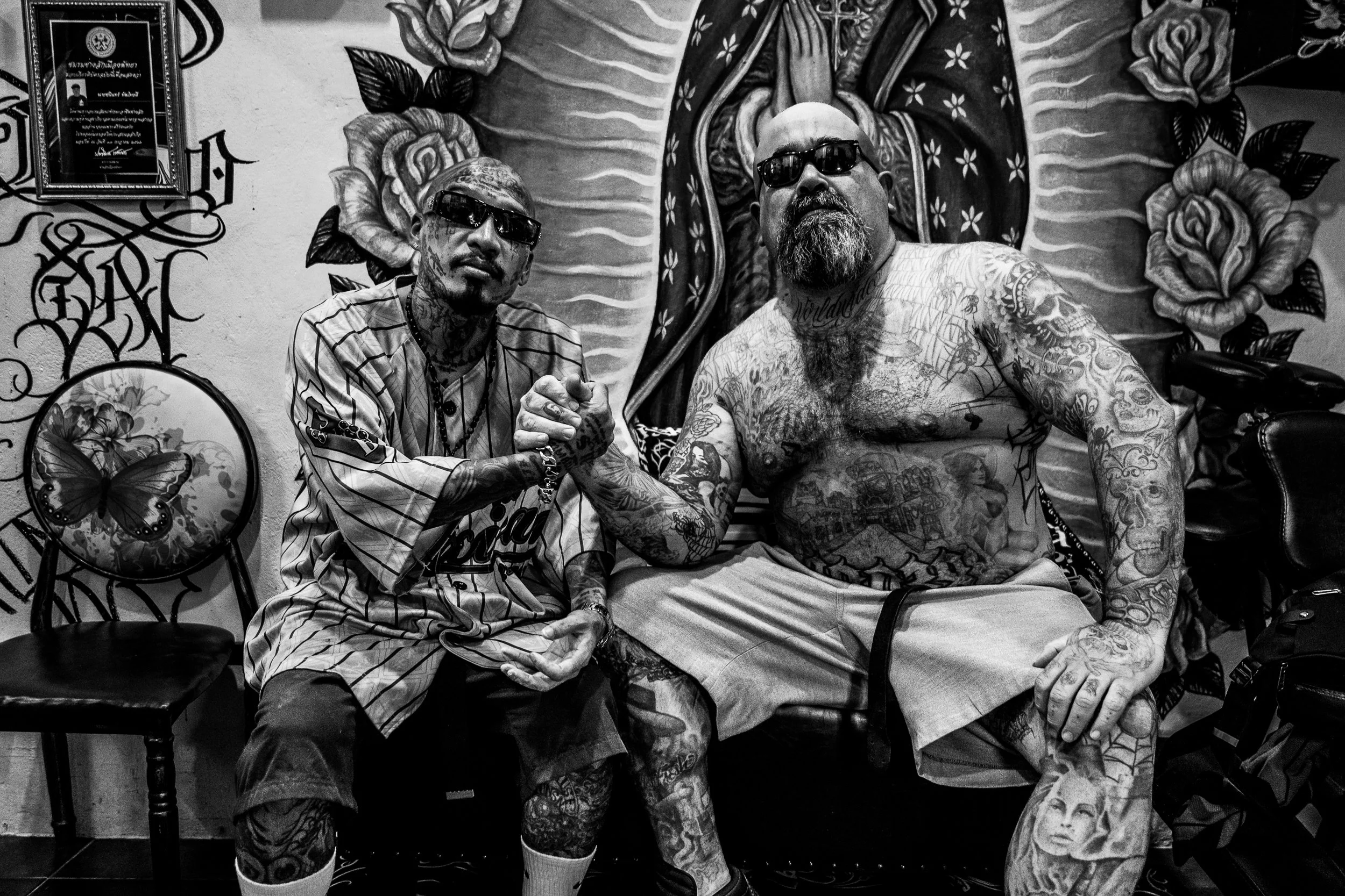 Two tattooed men, wearing sunglasses, engaging in a handshake in a room with floral and butterfly wall art, a framed certificate, and decorative murals.