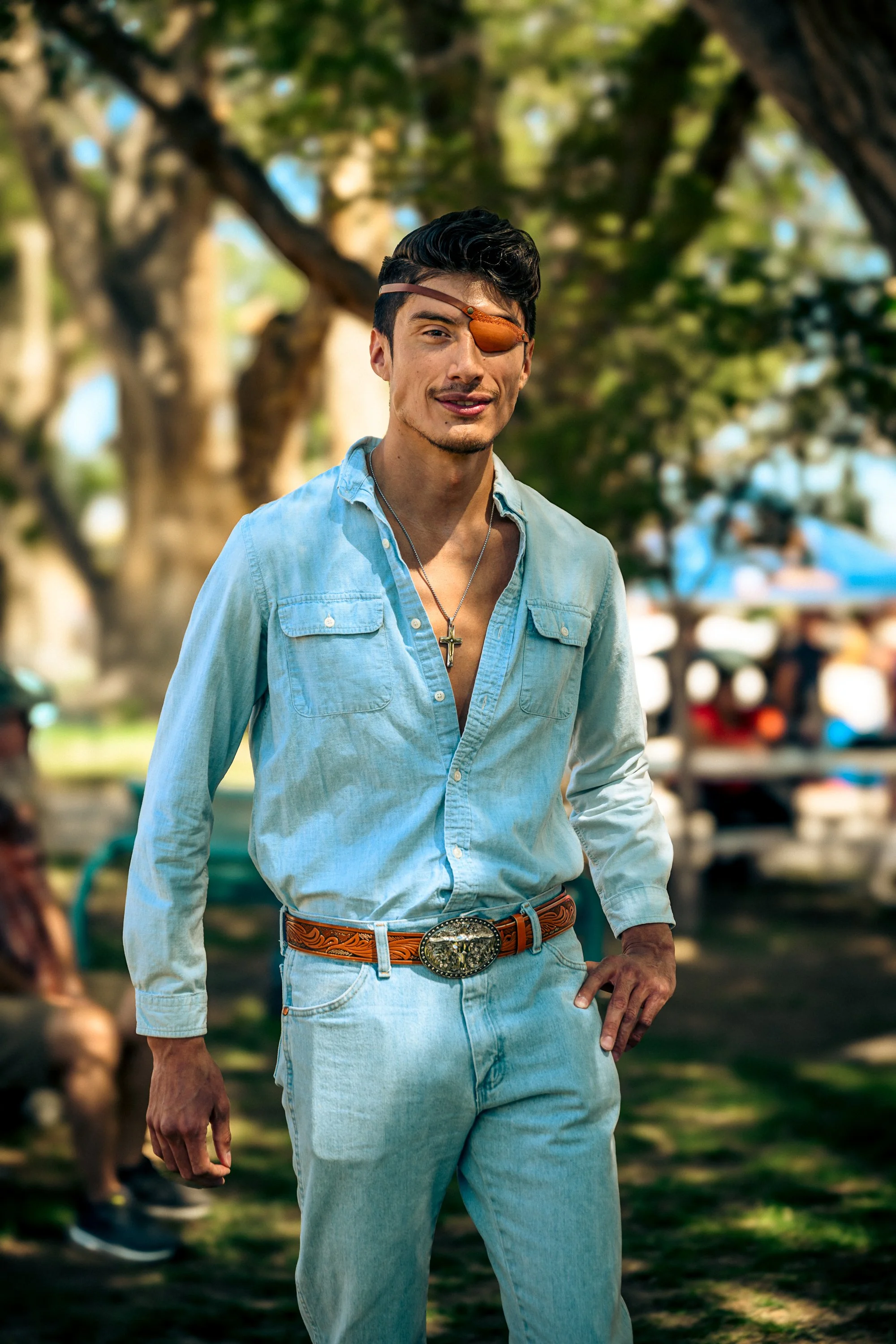 A man with short dark hair, wearing a light blue denim shirt partially unbuttoned, light blue jeans, an eye patch over his right eye, a necklace with a cross, and a large belt buckle, standing outdoors with trees and people in the background.