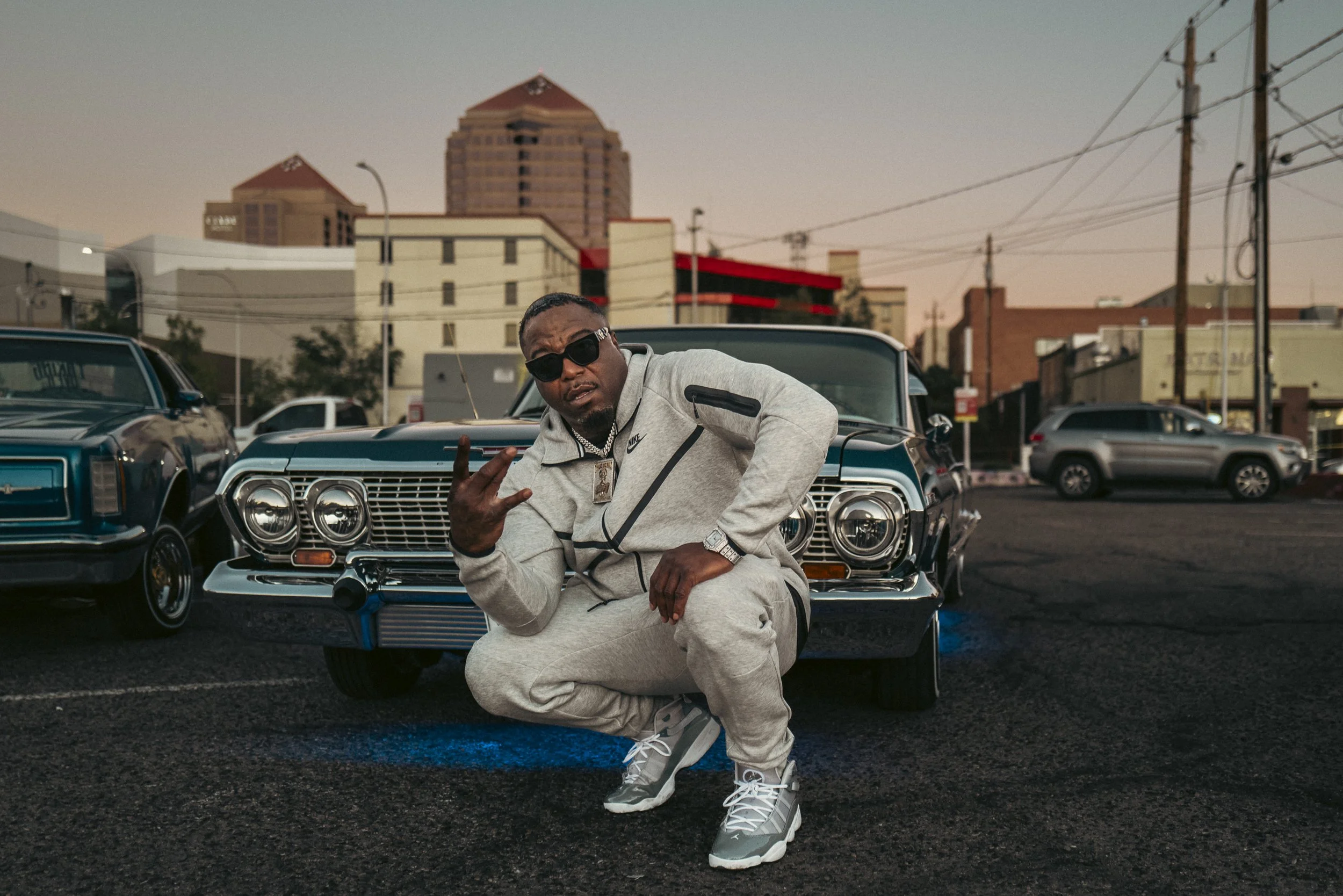 A man in gray athletic outfit squats in front of a black vintage car in an urban parking lot at dusk, making a hand gesture with his left hand and wearing sunglasses, jewelry, and sneakers.