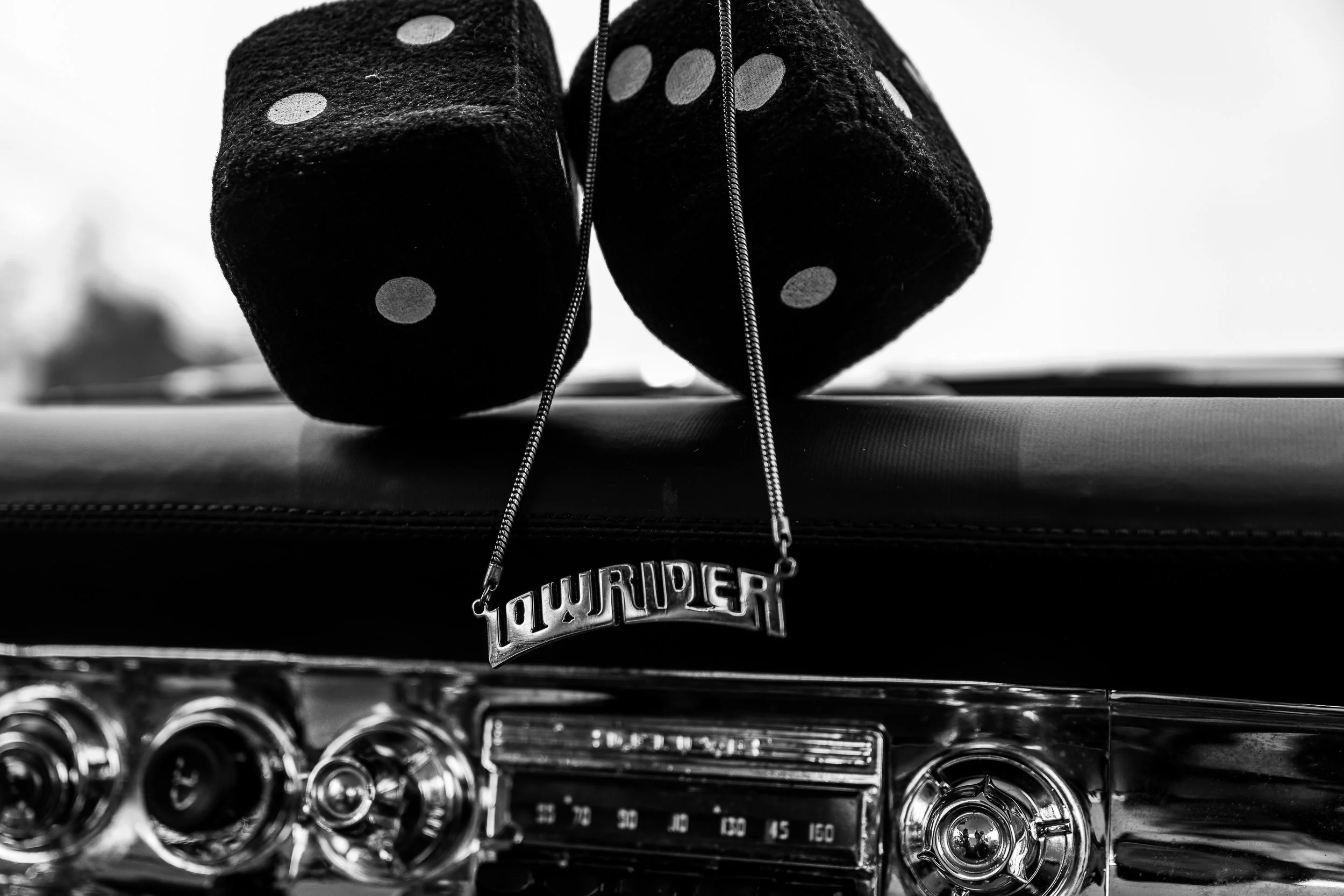 Black dice with white dots and a necklace with the word 'TOWRIDER' hanging from the dashboard of a vintage car, black and white photo.