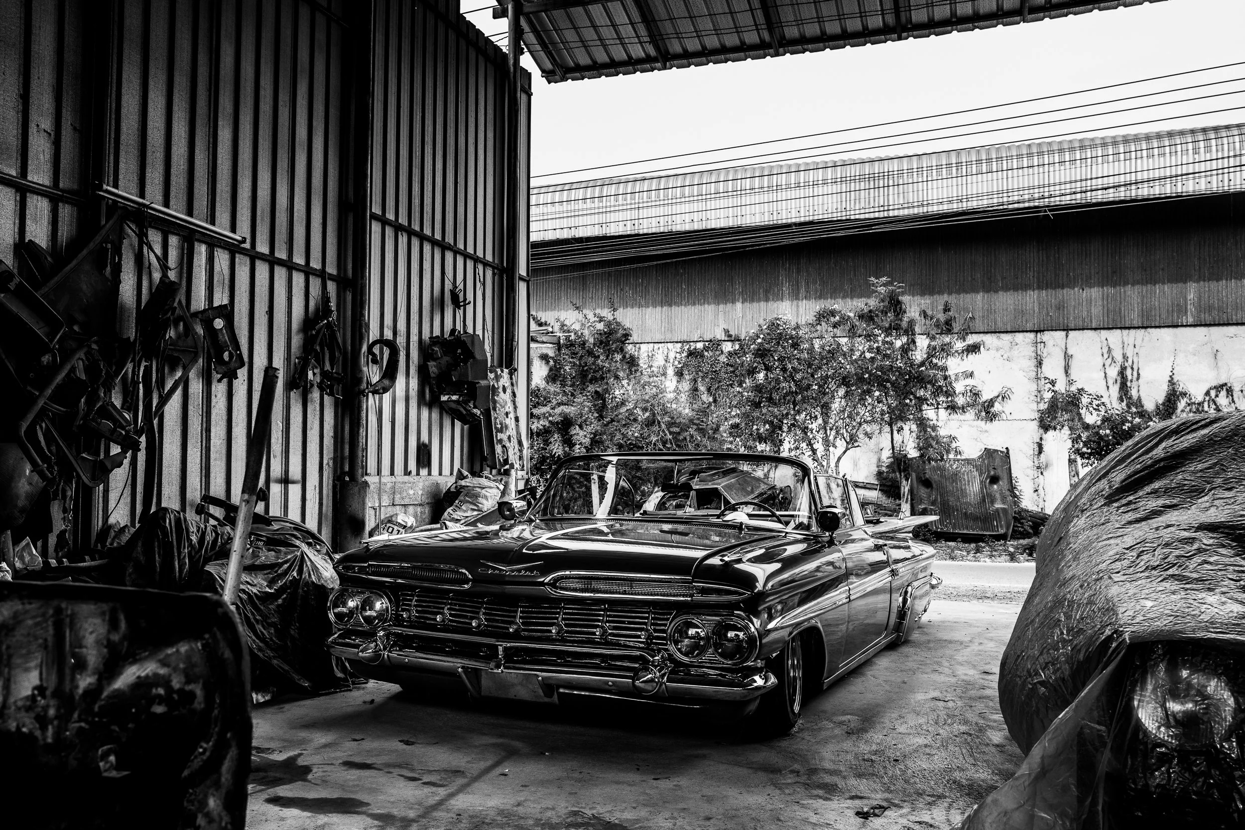 A black and white photo of a vintage car parked inside a cluttered garage or workshop with tools hanging on the wall and various objects and debris around.