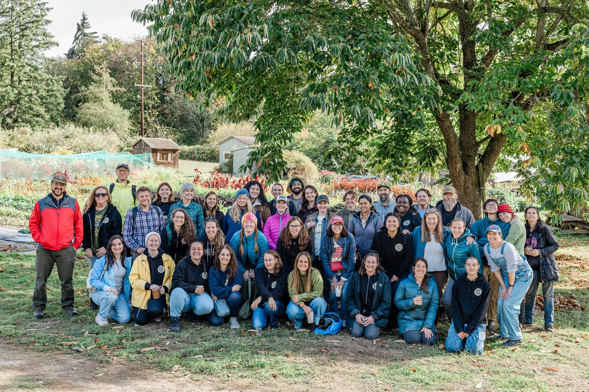 A large group of people posing outdoors under a tree in a garden or park setting.