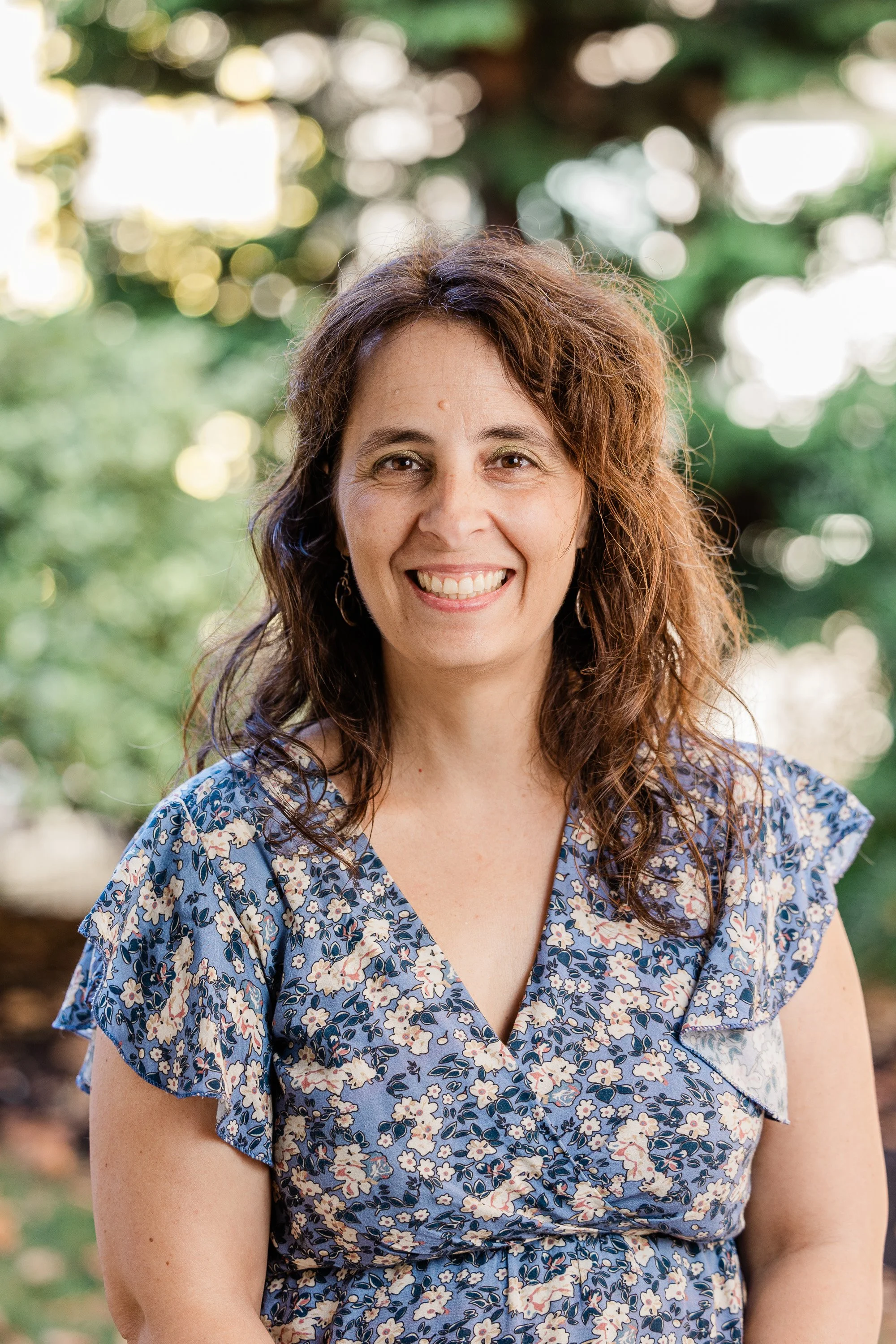 A woman with curly brown hair, wearing a purple sweater and earrings, smiling outdoors against a blurred dark background.