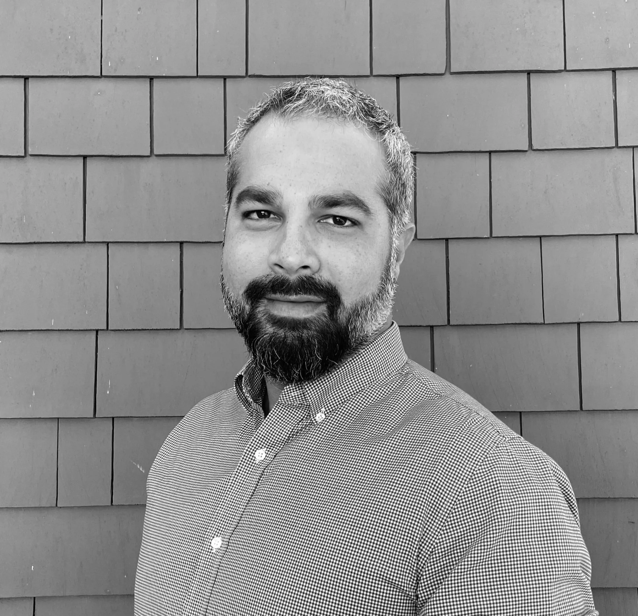 Black and white portrait of a man with a beard and short hair, wearing a button-up shirt, standing in front of a brick wall.