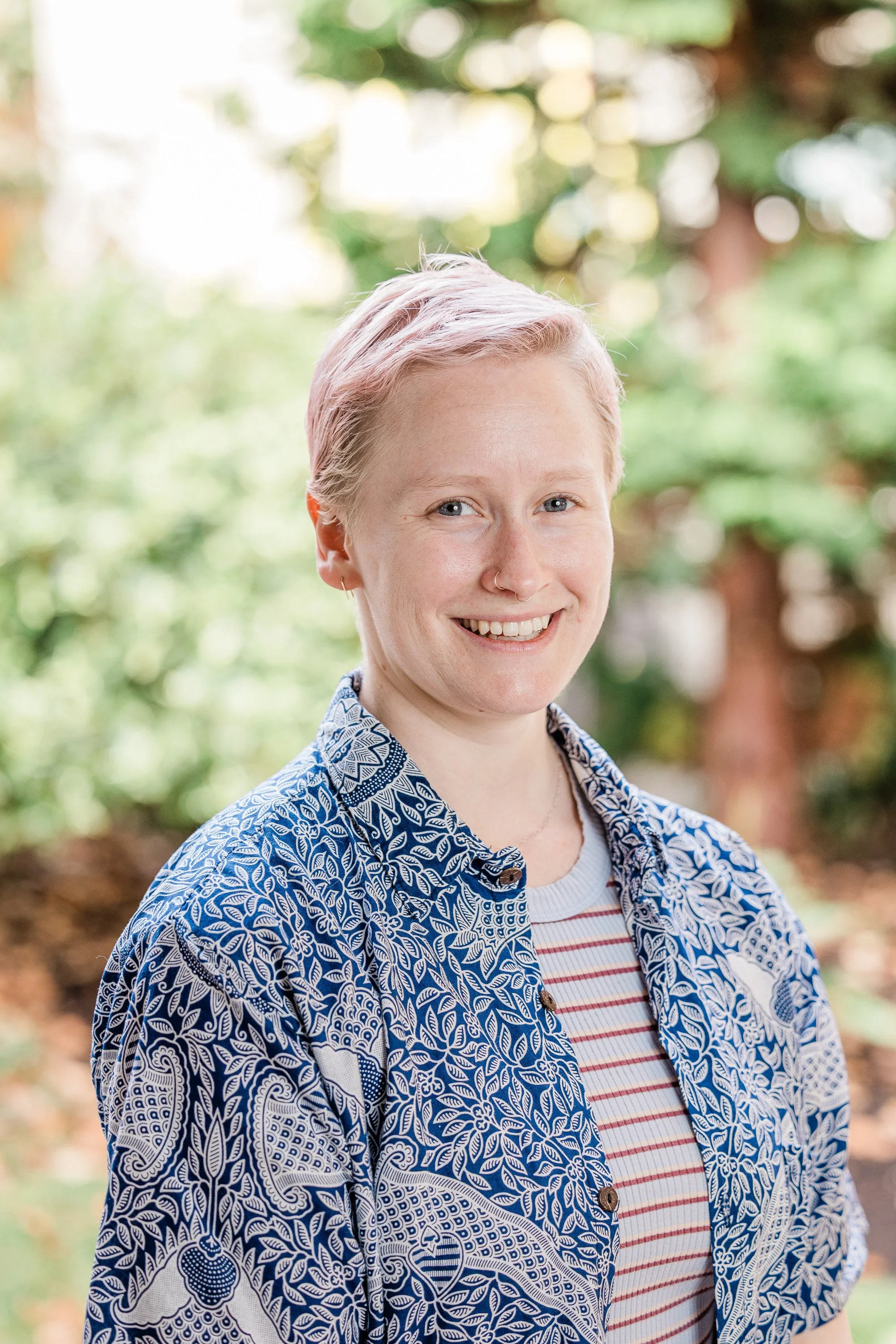 A woman with short pink hair and light skin, smiling, wearing a colorful patterned shirt, in an indoor setting.