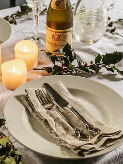 Cloth Napkins: Piglet in Bed's neutral napkin sits on a dinner plate accompanied by a fork and knife on top. The plate is on a white tablecloth surrounded by holly, two lit candles, and a wine.