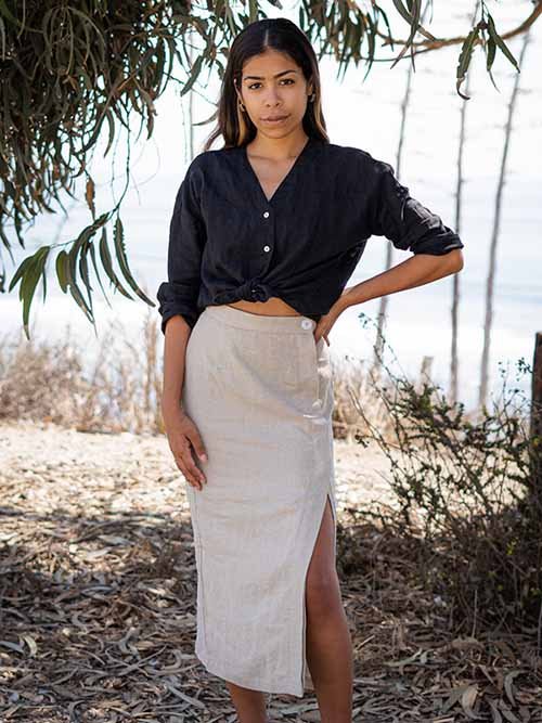 Eco-Friendly Clothing Brands: Model poses outside under some tree leaves wearing a black button down that's tied up in the front, on top of a neutral skirt with side slit.