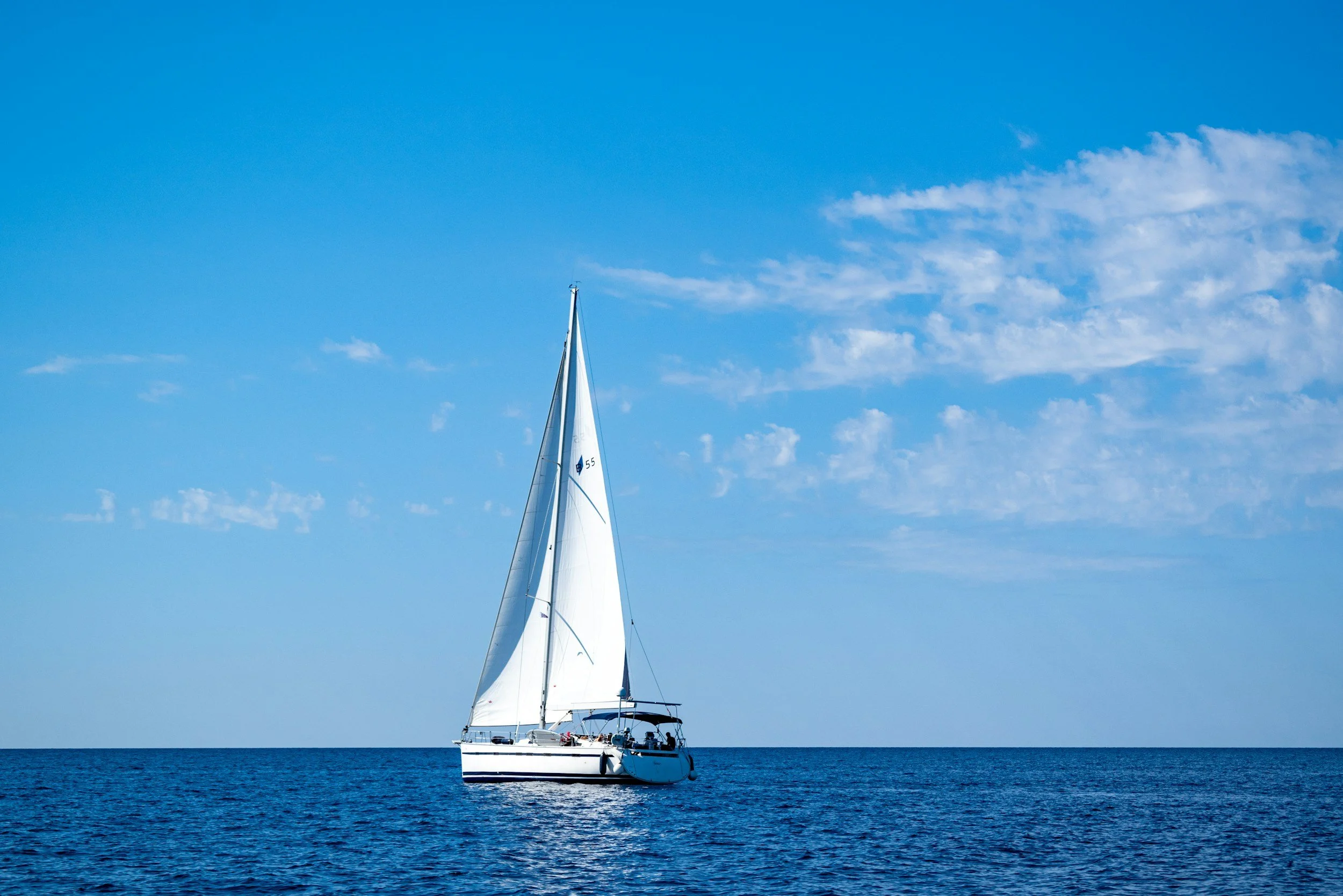 A white sailboat sailing on a calm blue ocean under a clear blue sky with a few wispy clouds.