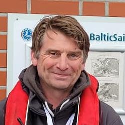 Man with light brown hair, wearing a black hoodie with a red and grey jacket, smiling outdoors in front of a sign that reads Baltic.