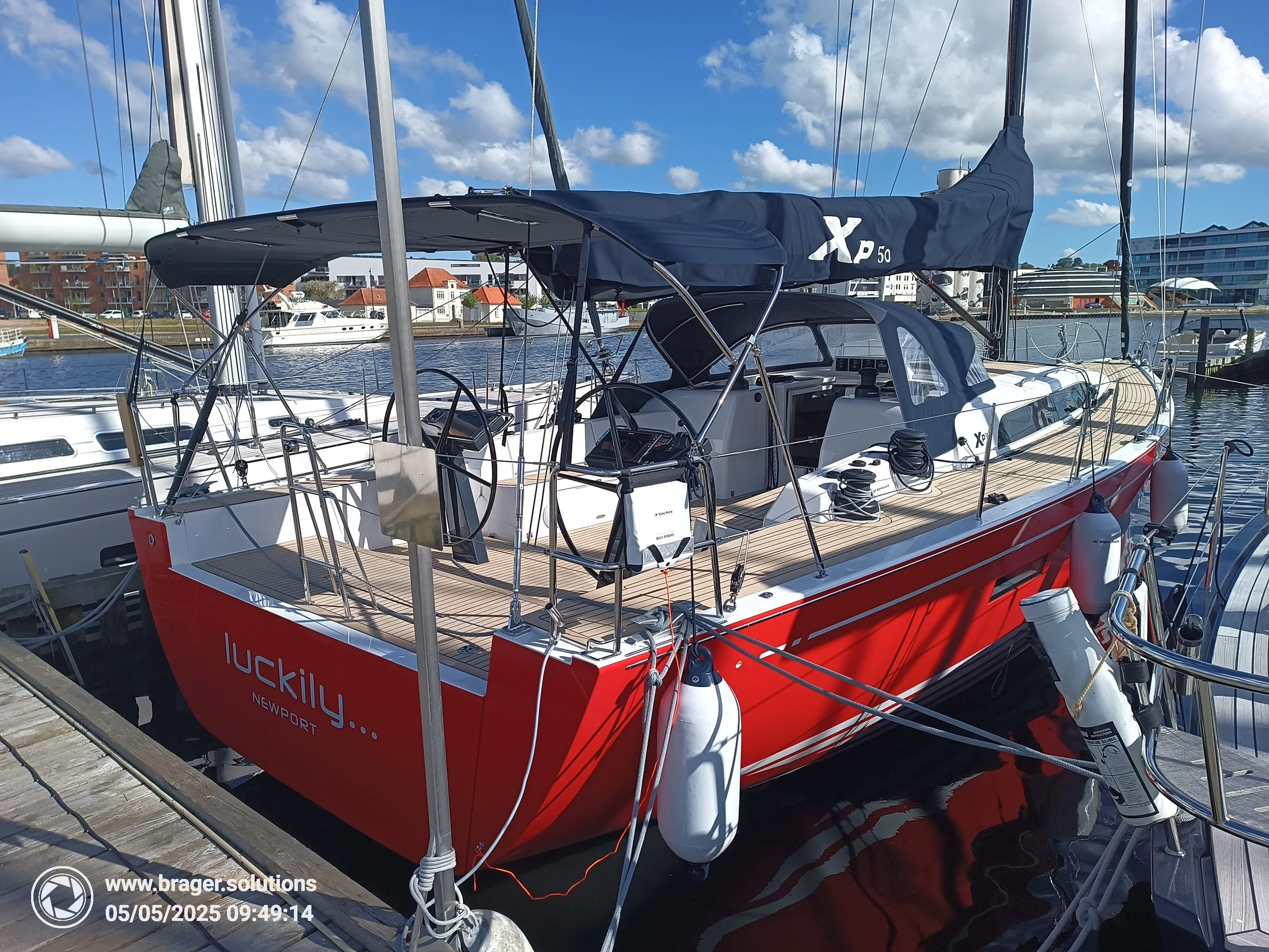 Red and white sailboat named 'Lucky' docked at a marina under a partly cloudy sky, with buildings and other boats visible in the background.