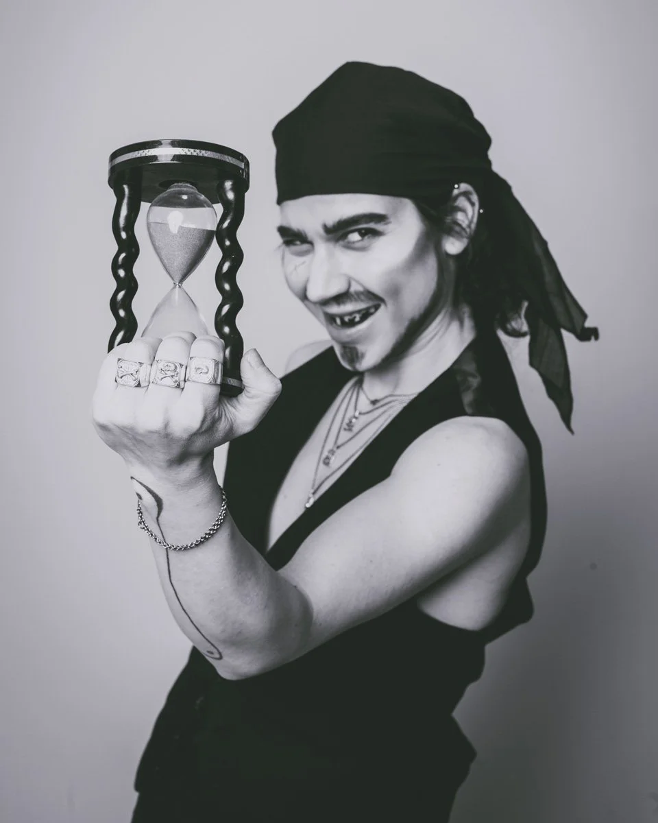 This image is of a drag king pirate holding an hourglass and wearing memento mori themed silver skull rings
