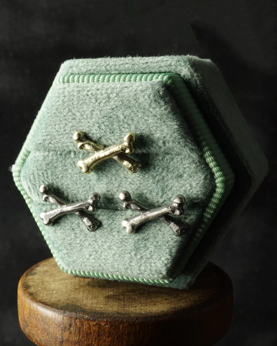 This image is of 3 small crossbone post earrings, 2 silver and one yellow gold, in a green jewelry box.