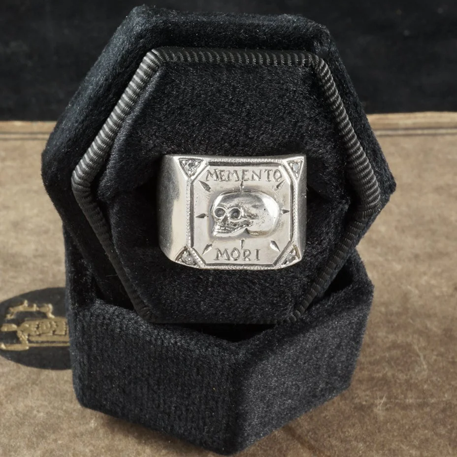 This image is of a silver skull ring with the words memento mori engraved, in a black ring box