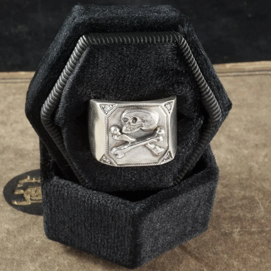 This image is of a skull and crossbones ring with hand engraved detail and salt and pepper diamond accent stones 
