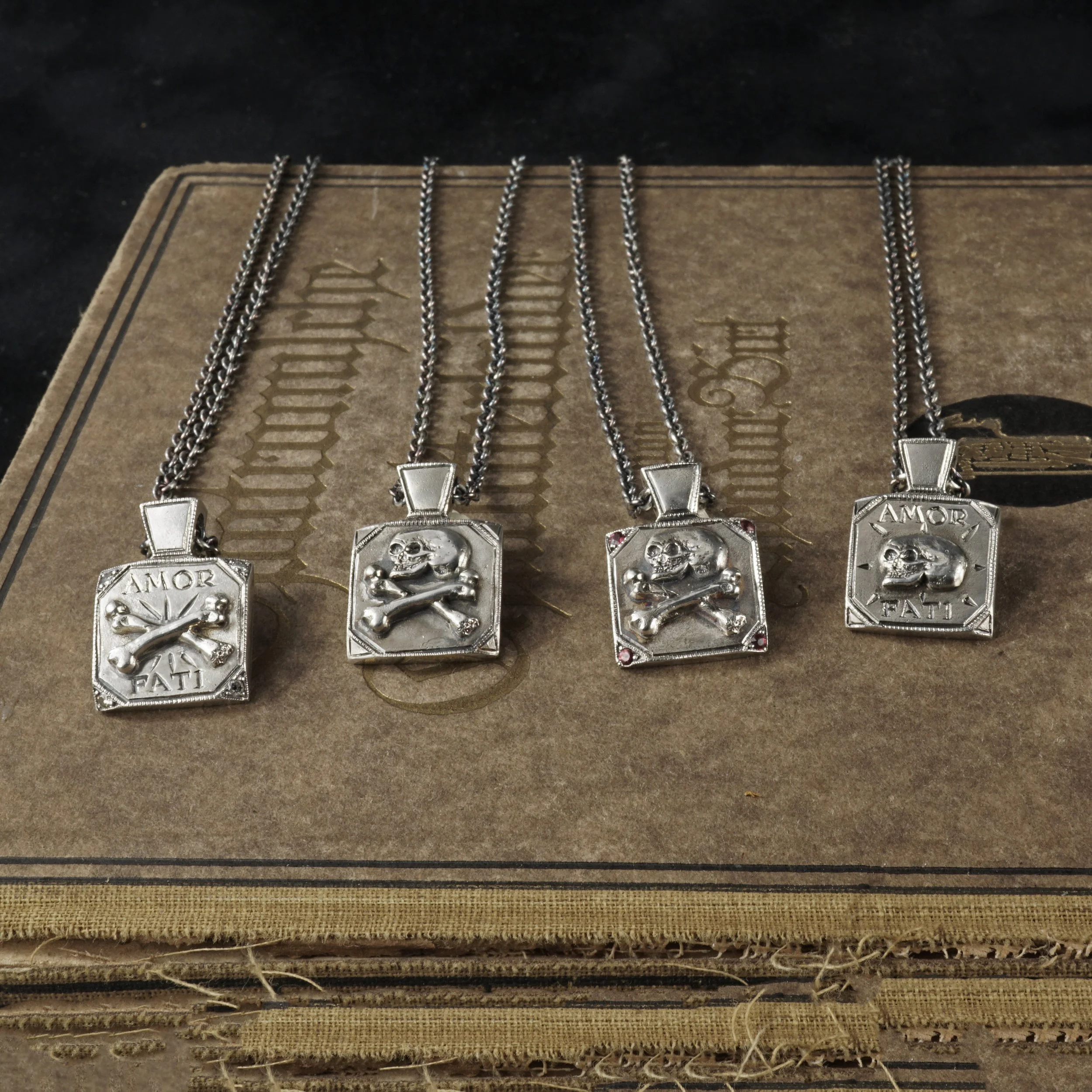 This image is of 4 silver skull pendants, 2 are skull and crossbones, and 2 have words memento mori engraved, sitting on a brown book