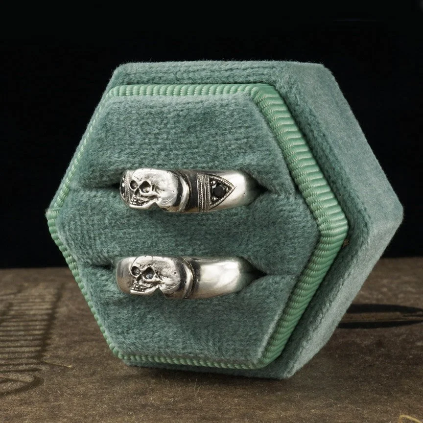 This image is of 2 small silver skull rings, one with black diamonds, in a green ring box