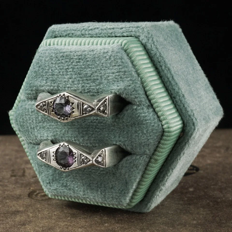 This image is of two silver rings, hand engraved, with purple spinel center stones and diamond accent stones, in a green ring box