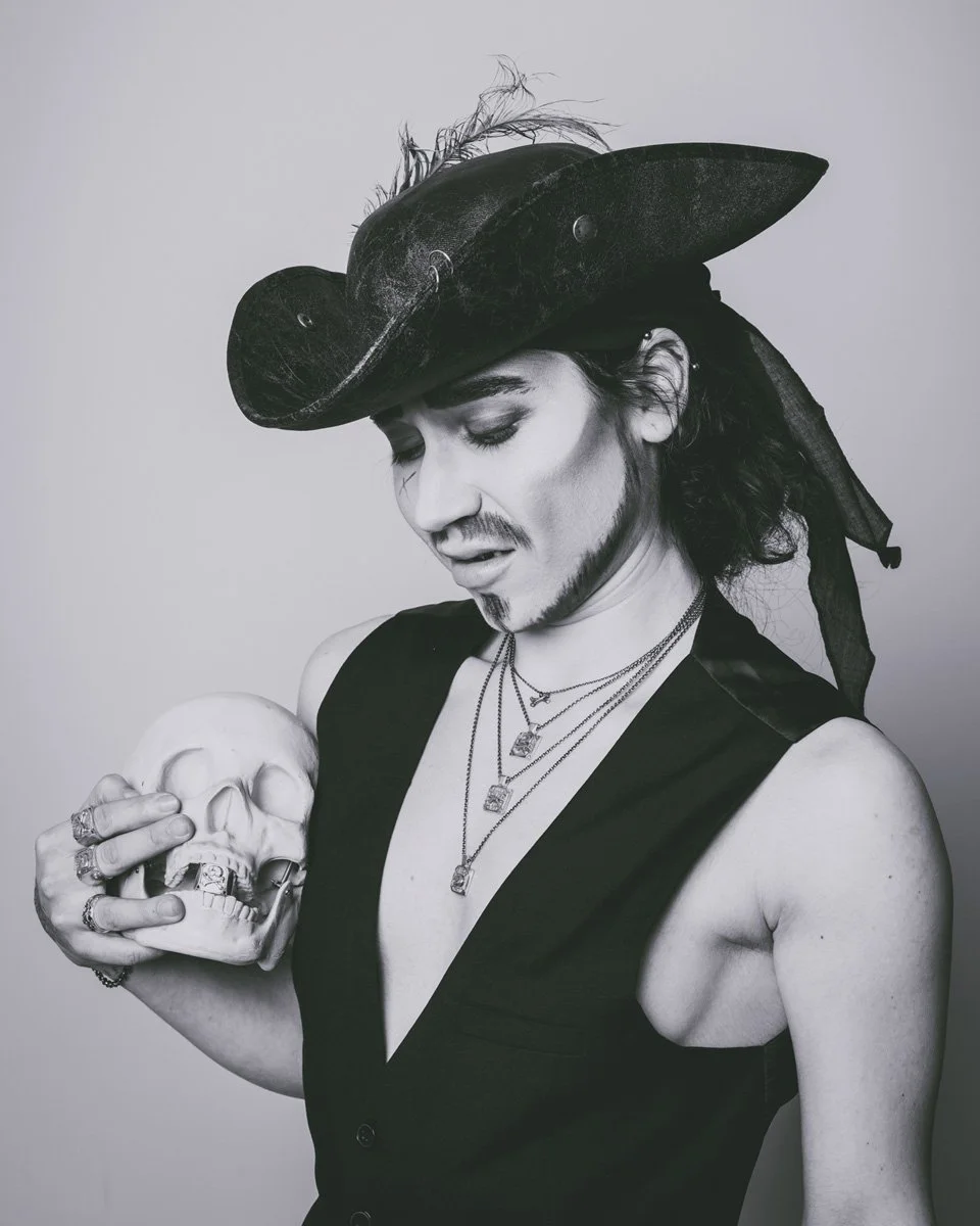This image is of a drag king pirate holding a plastic skull and wearing multiple pieces of skull and memento mori themed jewelry. Made by Anna Butwell for Metal Atelier in Berlin, Germany