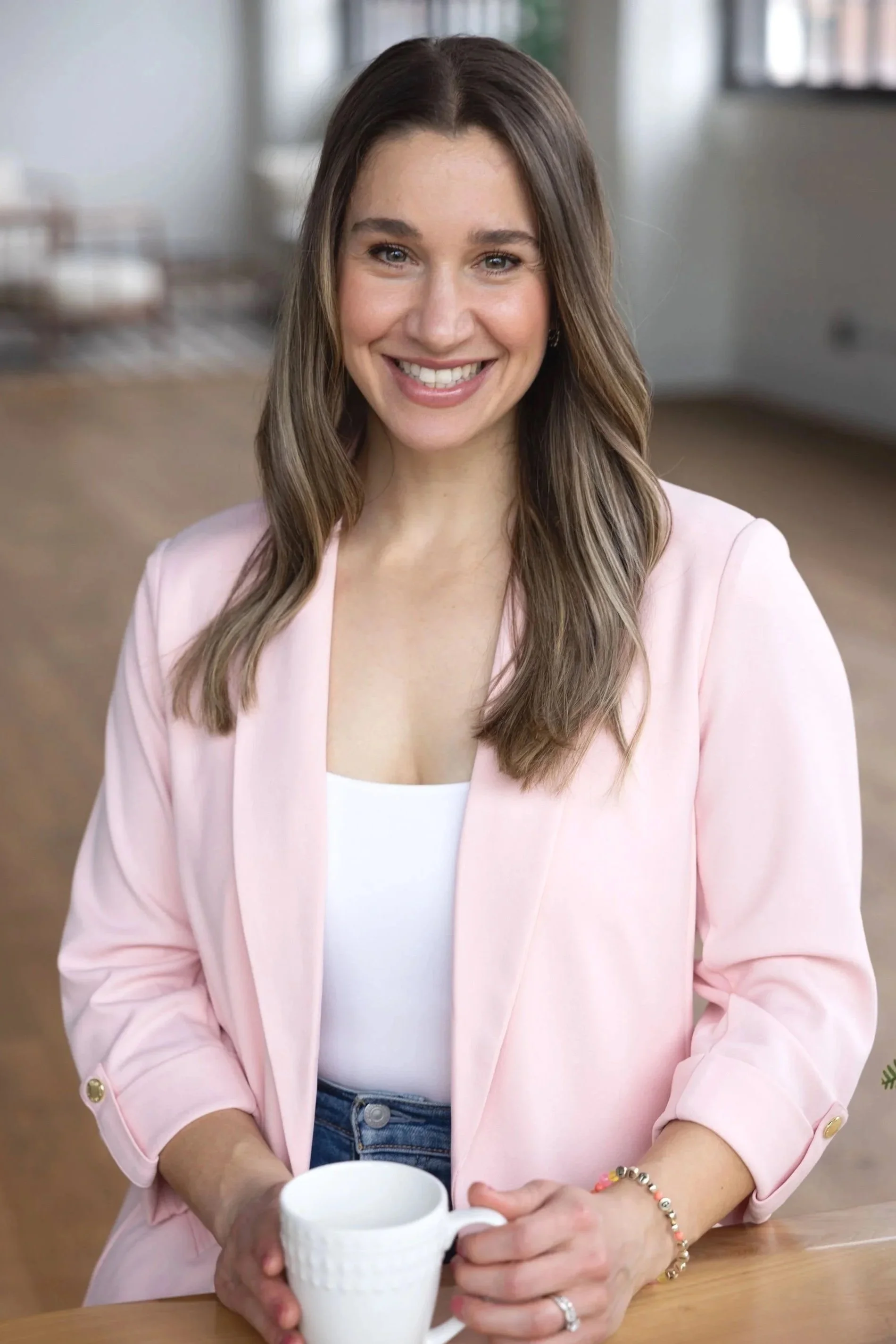 A woman with long brown hair, smiling naturally.  She is wearing a light pink blazer over a white top, with a beaded bracelet on her wrist and a ring on her finger. The background is in Cleveland Ohio. Branding photography near me.