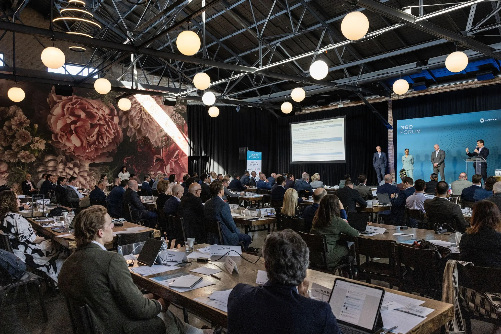 Attendees seated at long tables during a corporate conference session in an industrial event venue with globe lights