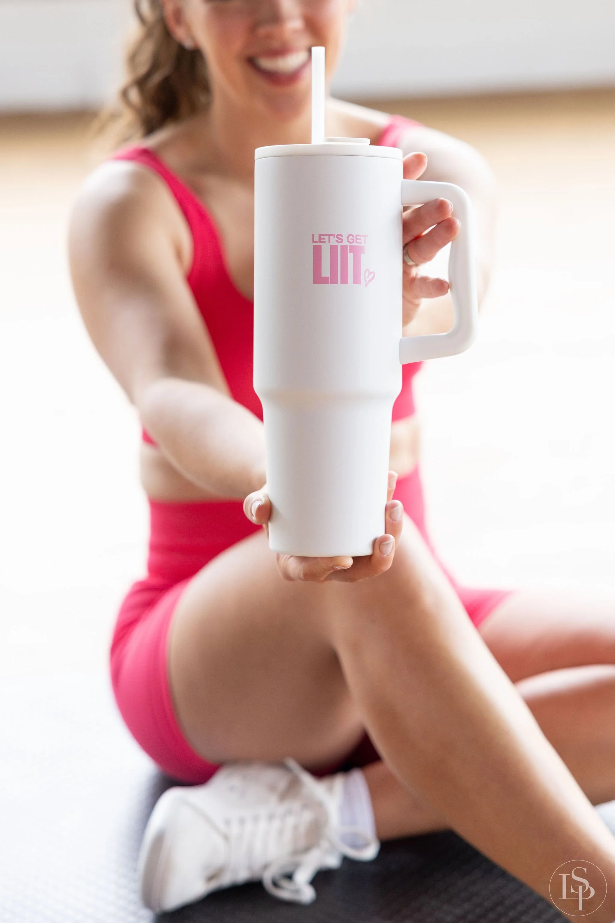 Social Media Content photographer near me. Woman in pink athletic wear sitting on the floor, holding a white tumbler with a straw, smiling. Serving greater Cleveland & Akron Ohio areas.