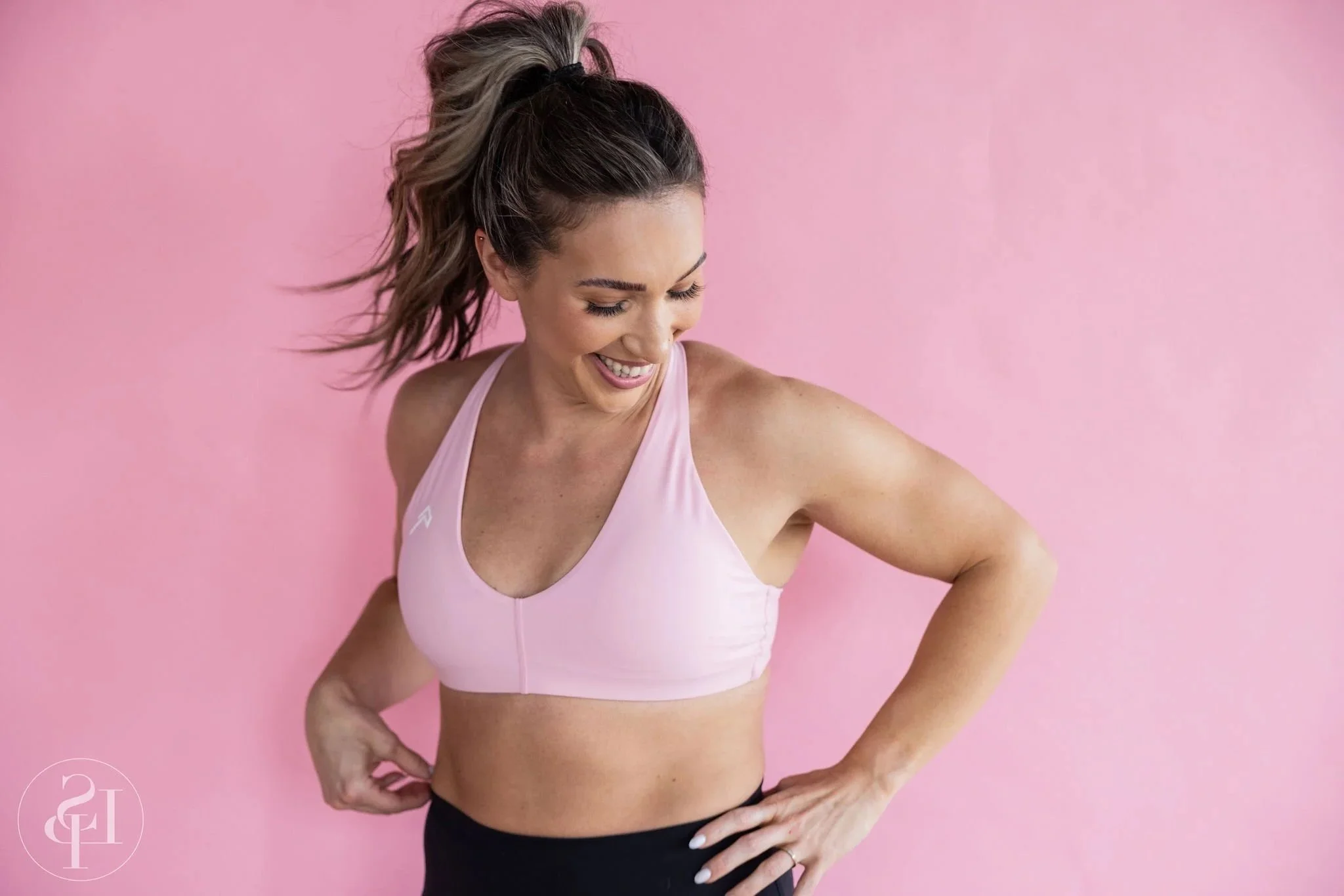 A woman in athletic attire with a pink background, smiling and looking down, with her hands on her hips. Commercial brand photographer near me in Cleveland or Akron Ohio. Richfield, Bath, Independence, Hudson, luxury experience.