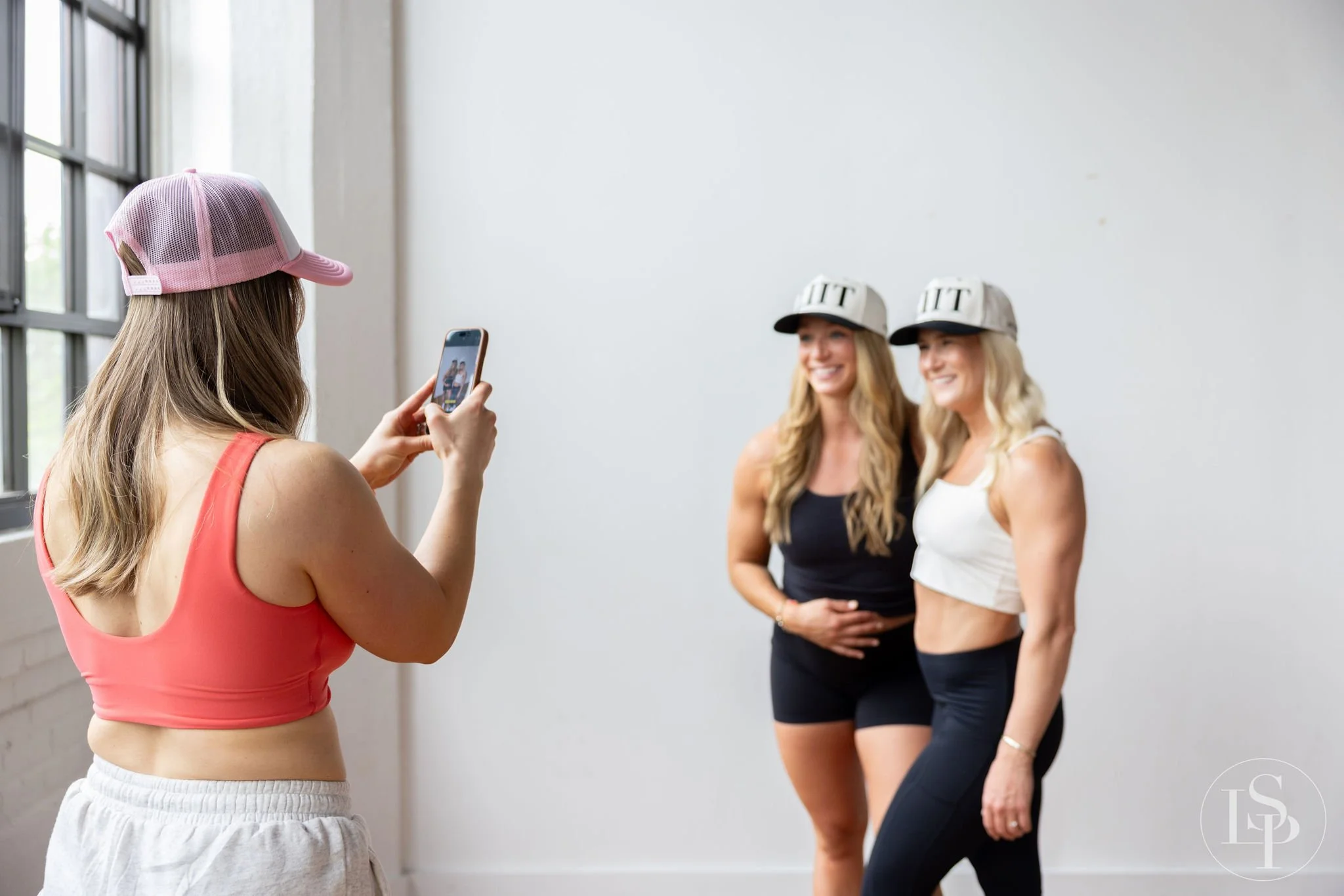 A woman with long blonde hair wearing a pink tank top and pink cap takes a photo of two women with long blonde hair wearing matching gray and black caps and black and white workout clothes, standing against a white wall in a studio.