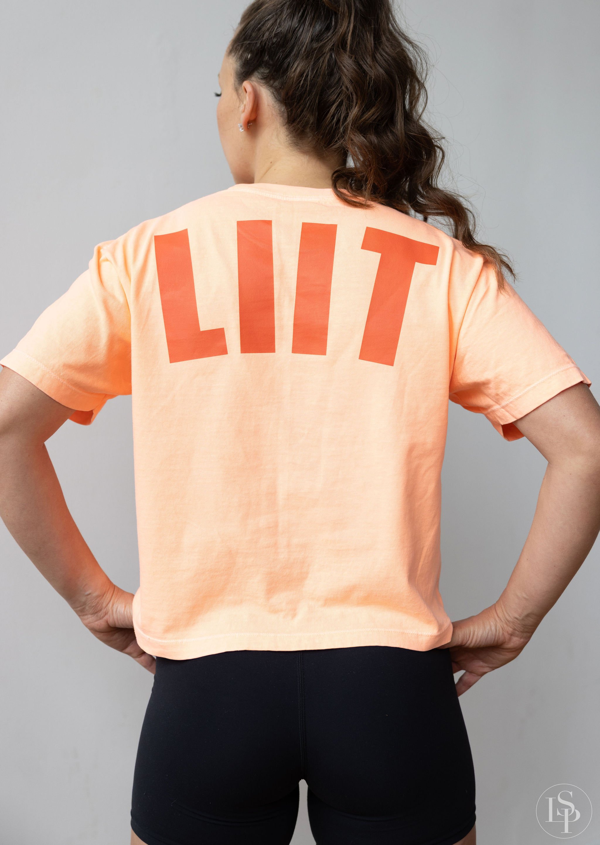 Back view of woman with curly brown hair wearing a peach T-shirt with red letters spelling 'LIT' on the back, and black shorts, standing against a gray background. Commercial brand photographer for health and wellness industry. Serving Cleveland