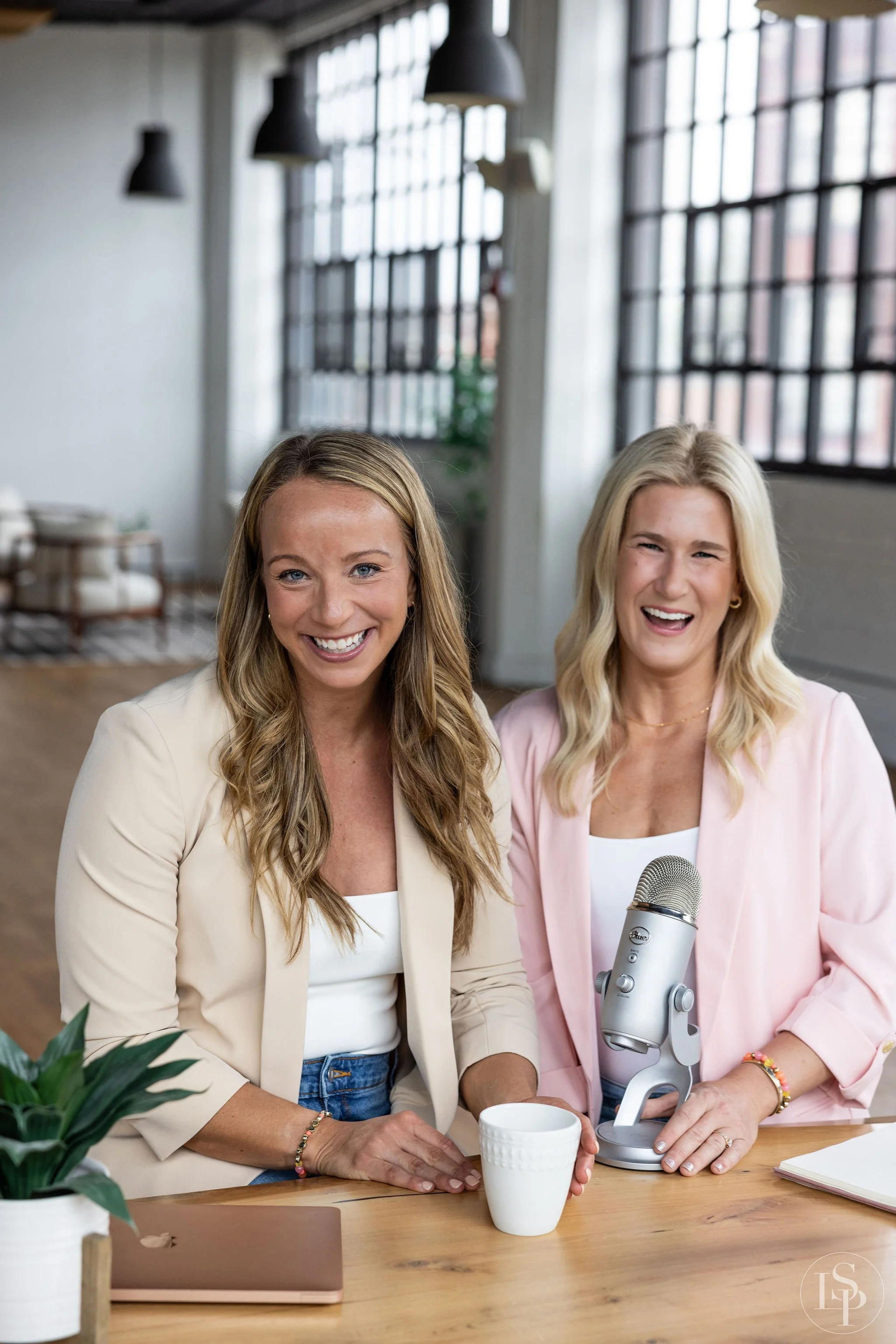 A lifestyle team portrait of Two women sitting at a table in a bright, modern office space, smiling at the camera; one woman is holding a microphone, and there is a mug, a laptop, and a plant on the table. Brand photographer near me in Cleveland.