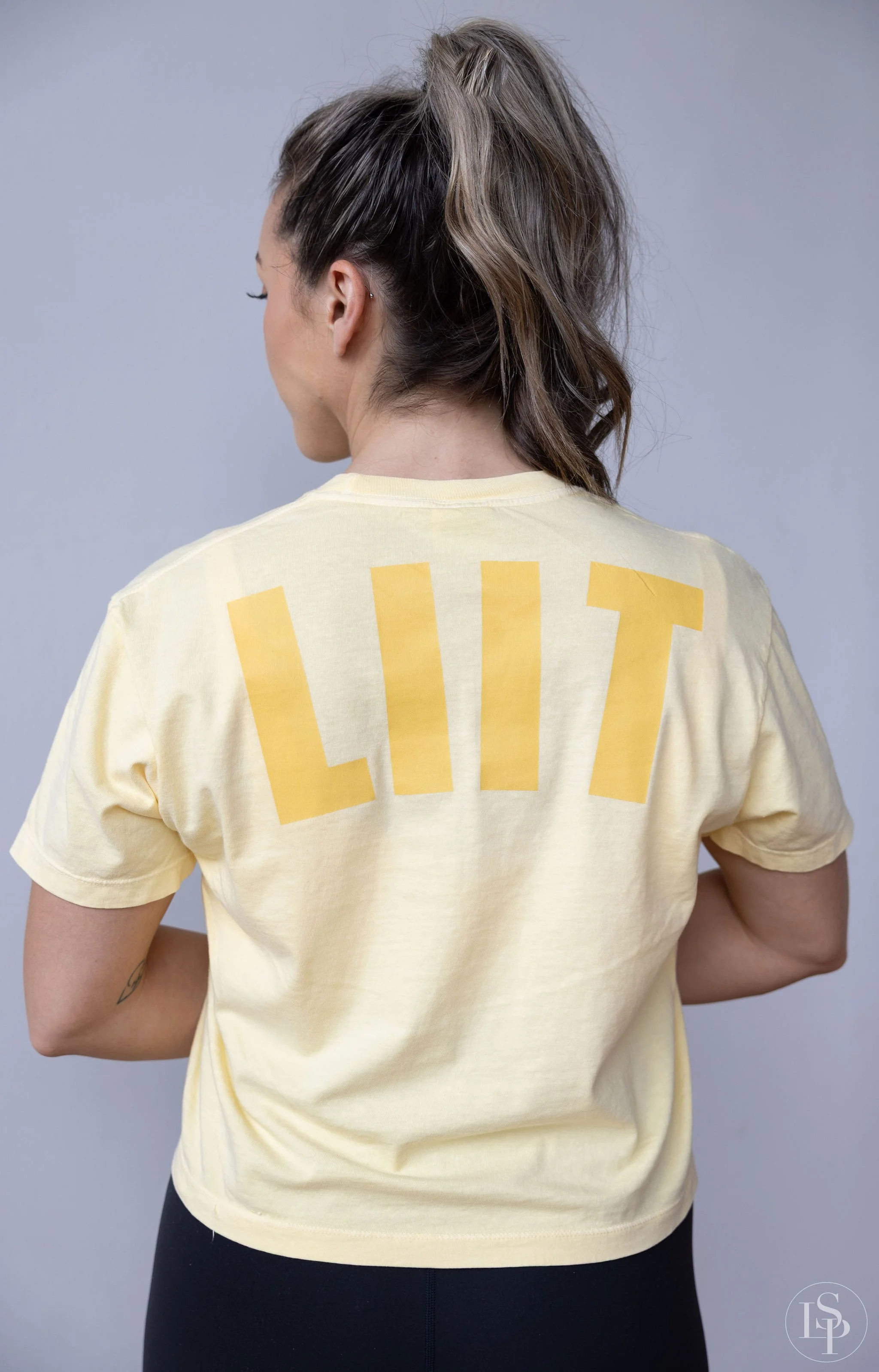 Back view of a woman wearing a yellow t-shirt with the letters 'LIT' printed in large yellow font. She has wavy brown hair tied in a high ponytail and is standing against a plain gray background. Commercial brand photographer near me in Cleveland.