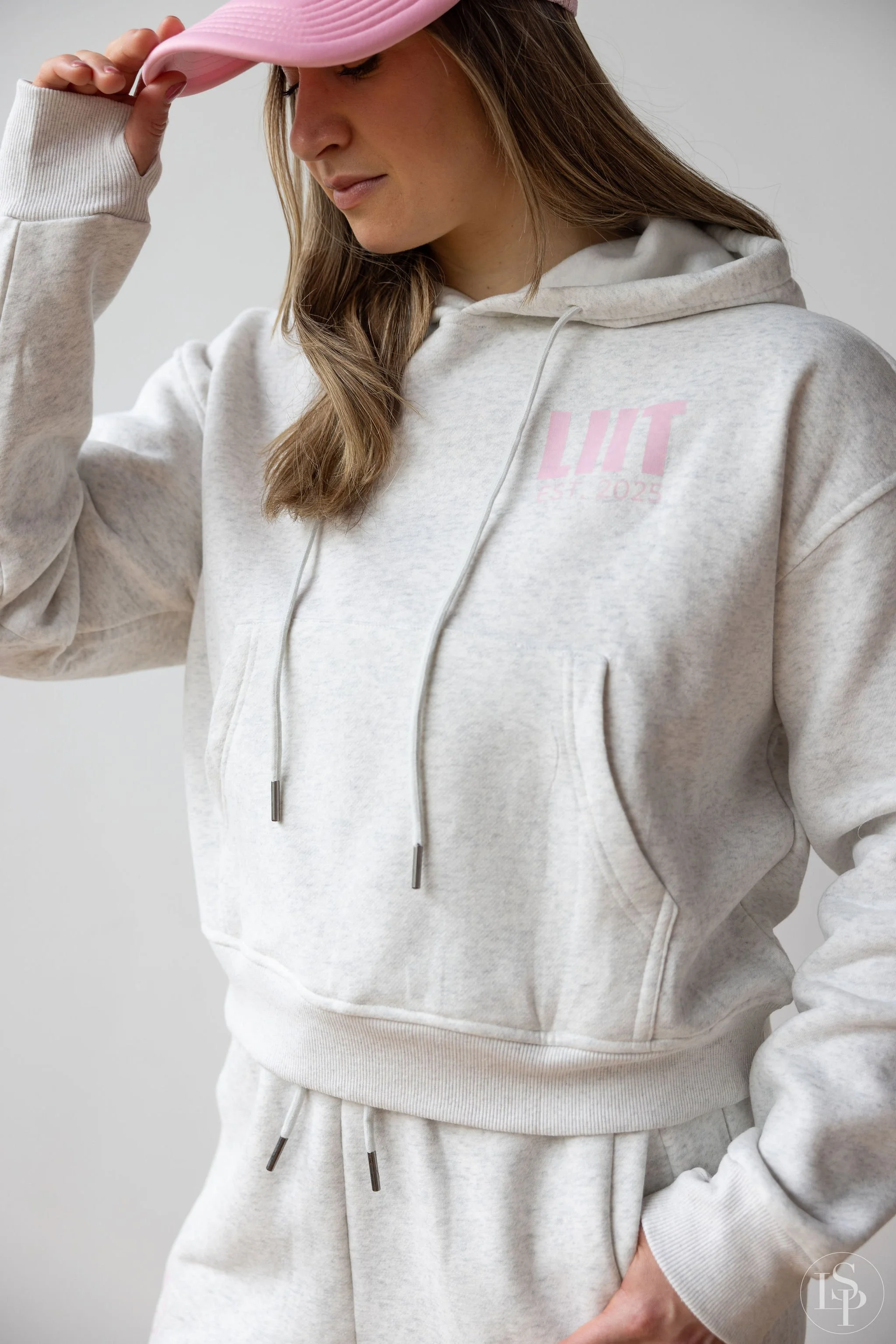 A woman wearing a light gray hoodie and sweatpants, holding a light pink cap, standing against a plain light background. Editorial fashion and commercial photographer near me in cleveland or Akron ohio.