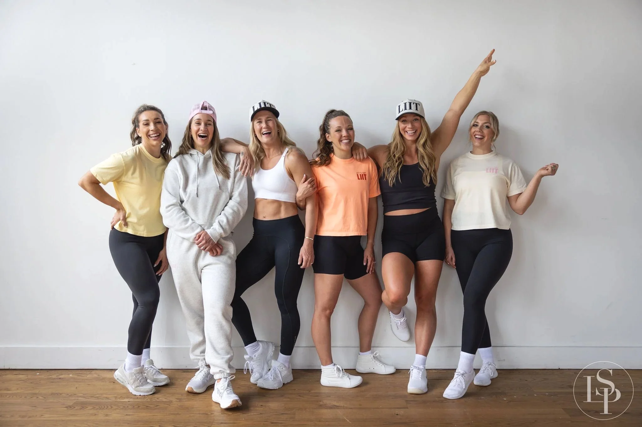 Group headshot near me of six smiling women standing against a white wall, dressed in athletic wear, with arm around each other, some wearing baseball caps and sneakers. Taken in Cleveland Ohio.