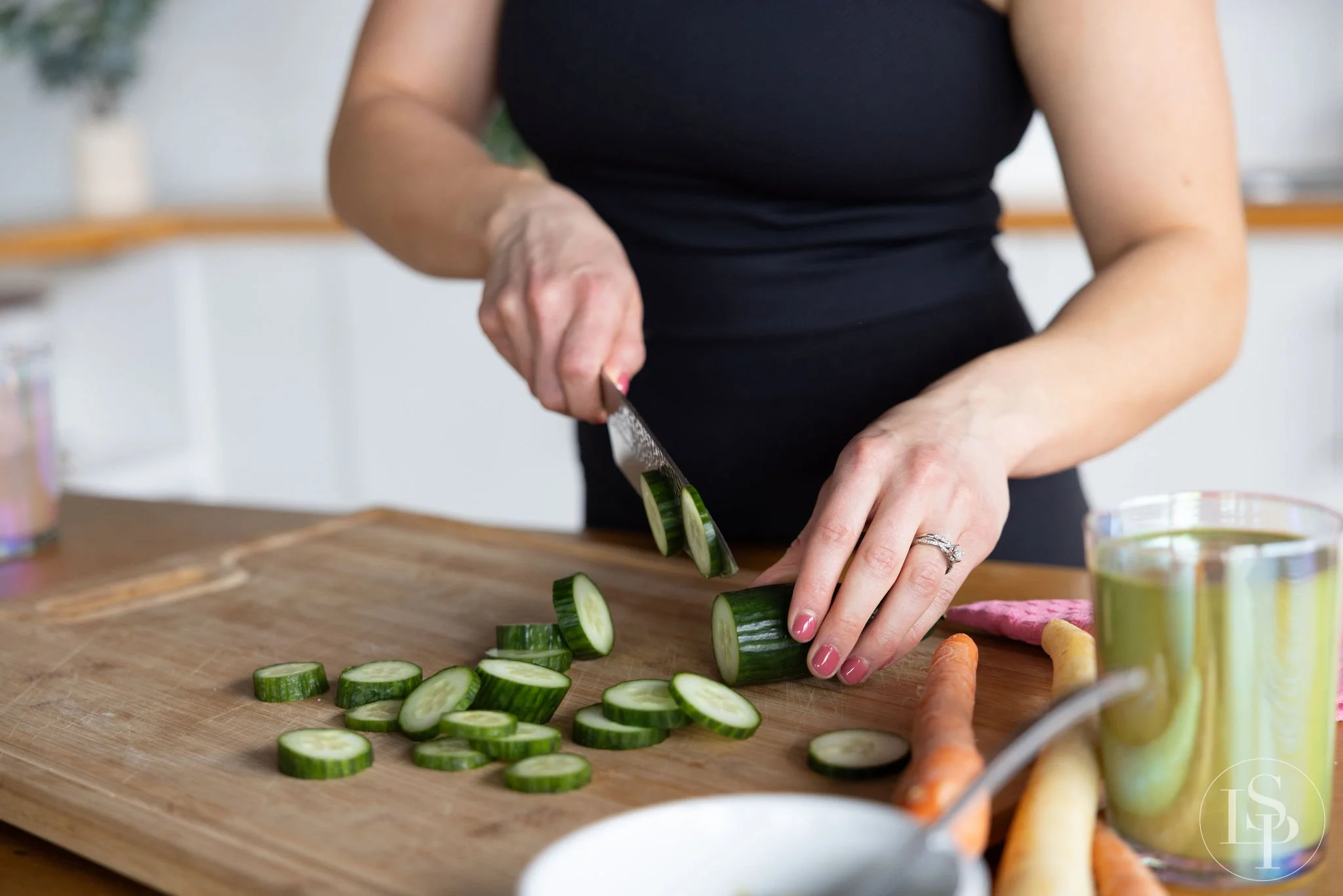 A person is slicing cucumbers on a wooden cutting board in a kitchen. There are carrots and various drinks nearby.