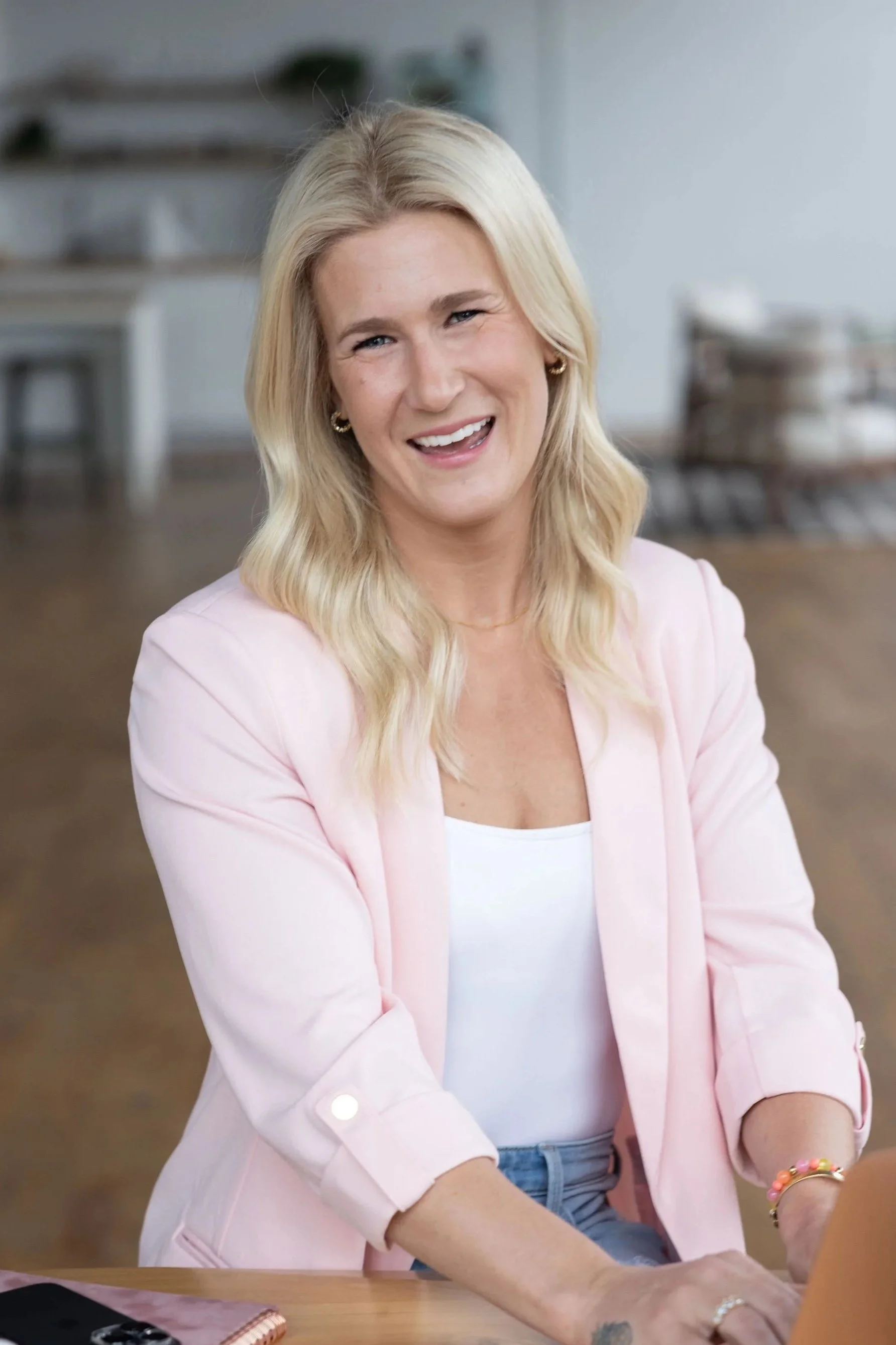 A woman with blonde hair, wearing a pink blazer and gold hoop earrings, smiling and engaging in a conversation in a bright, modern room.