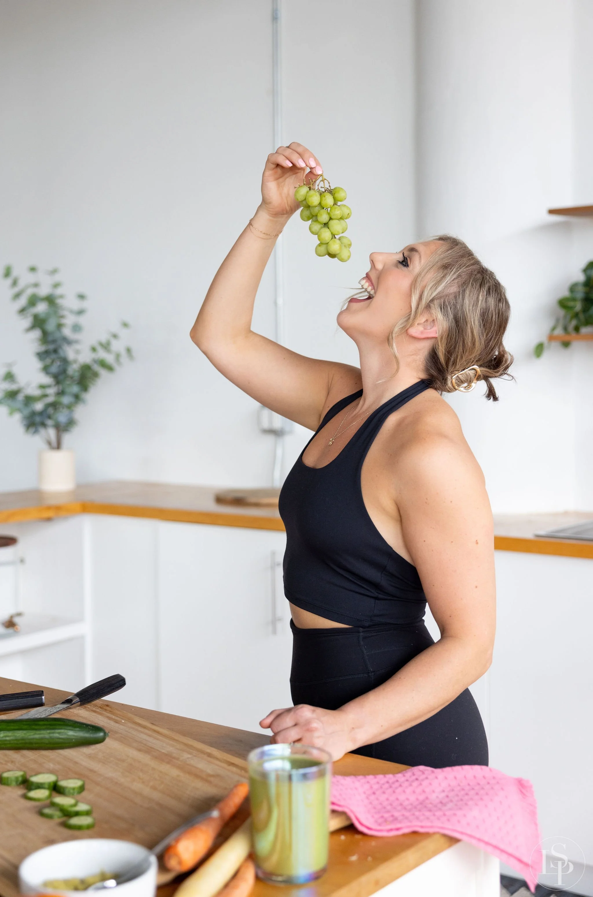 A woman with green grapes above her open mouth in a bright kitchen. The kitchen has white cabinets, and there are cucumbers, carrots, a pink towel in health and wellness brand photographer near me. Commercial brand photographer near me.