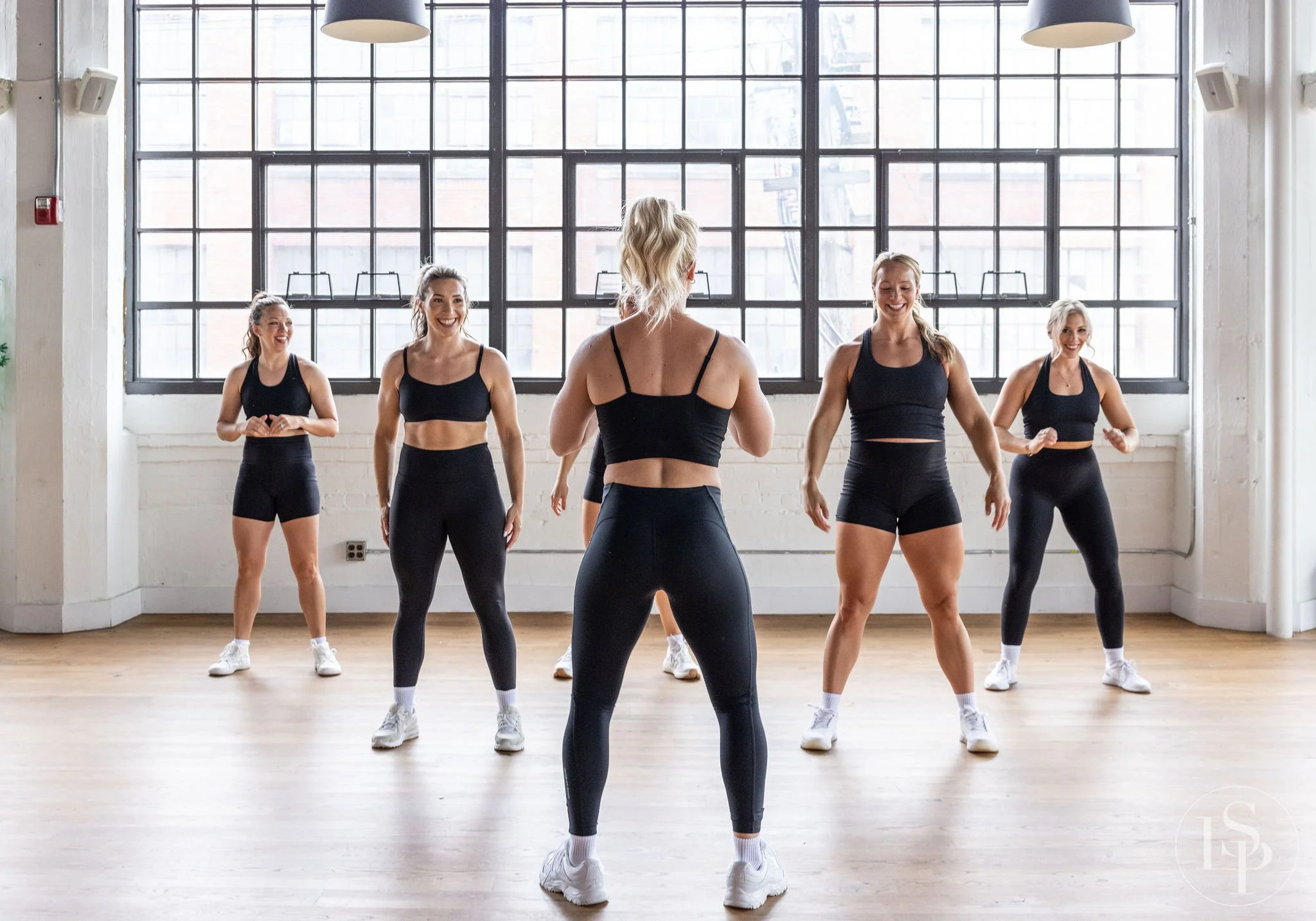 Group of women in workout clothes participating in a fitness class in a bright studio with large windows. Health and wellness branding photographer near me.