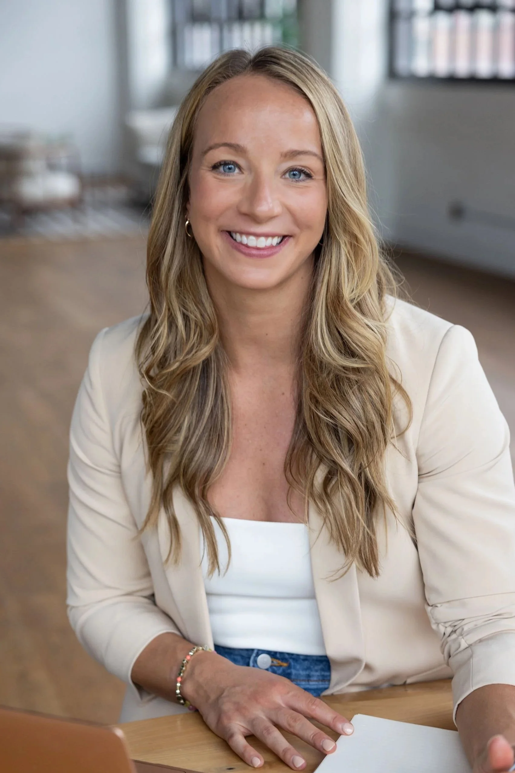 A professional lifestyle headshot of a woman with blonde hair, taken in Cleveland Ohio - smiling while sitting at a table with papers, wearing a white shirt and blazer in a bright room with large windows.